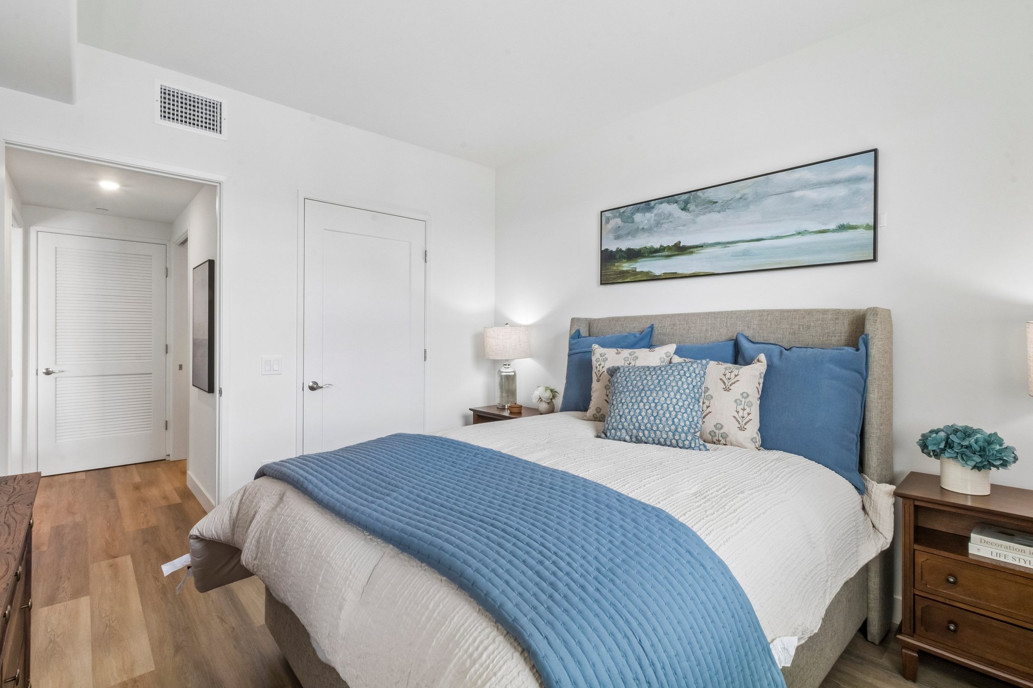 A modern bedroom featuring a queen-sized bed with a blue and white color scheme. The bed is adorned with decorative pillows and a pastel blue throw blanket. Two bedside lamps illuminate wooden nightstands, and a landscape painting hangs above the bed, complementing the serene decor. A doorway leads to a hallway.
