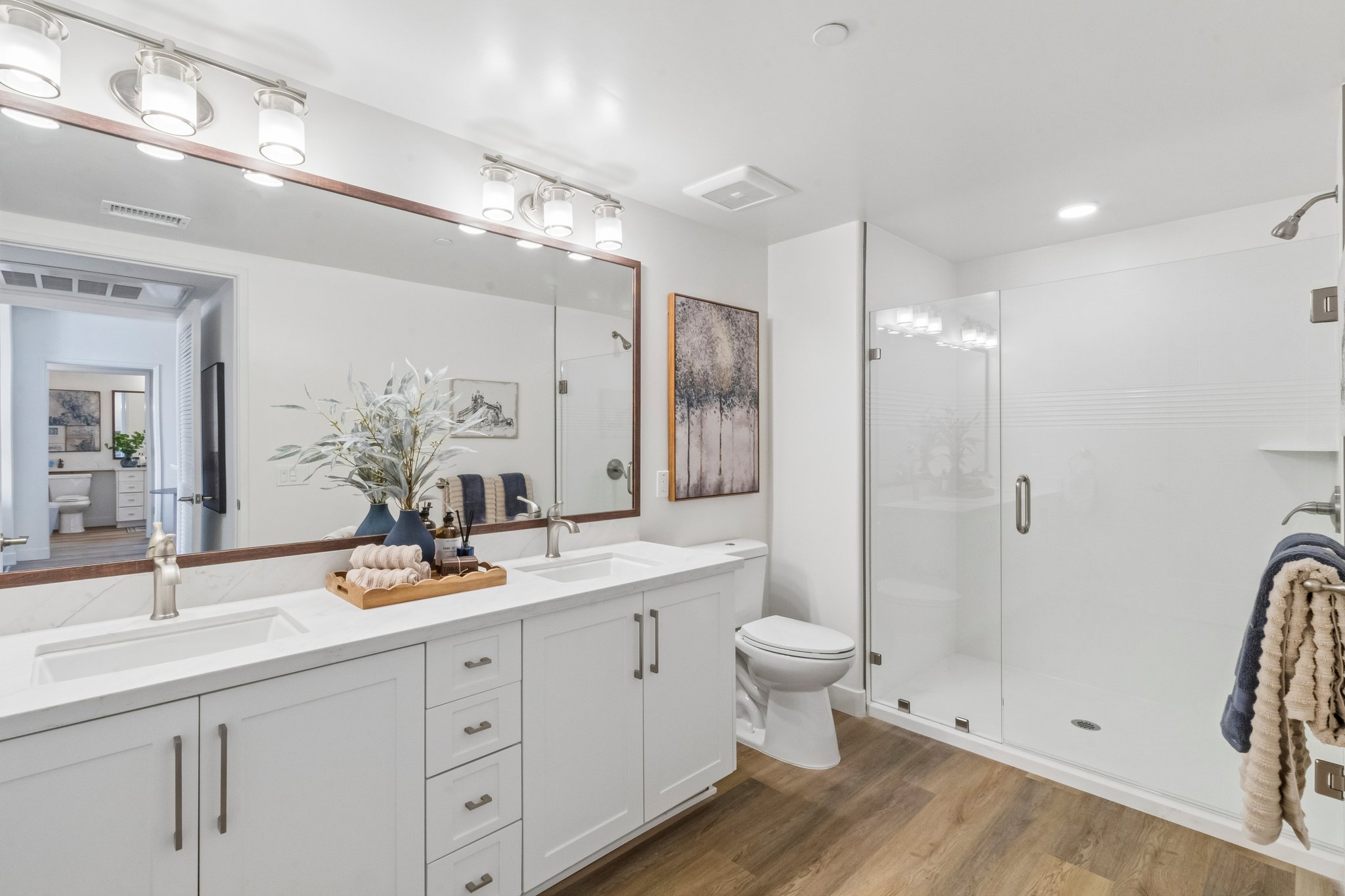 A modern bathroom featuring a double vanity with two sinks, large mirrors, stylish lighting fixtures, a glass shower enclosure, and a neutral color scheme. Decorative elements include a vase with greenery and neatly arranged towels. The floor is wooden, adding warmth to the space.