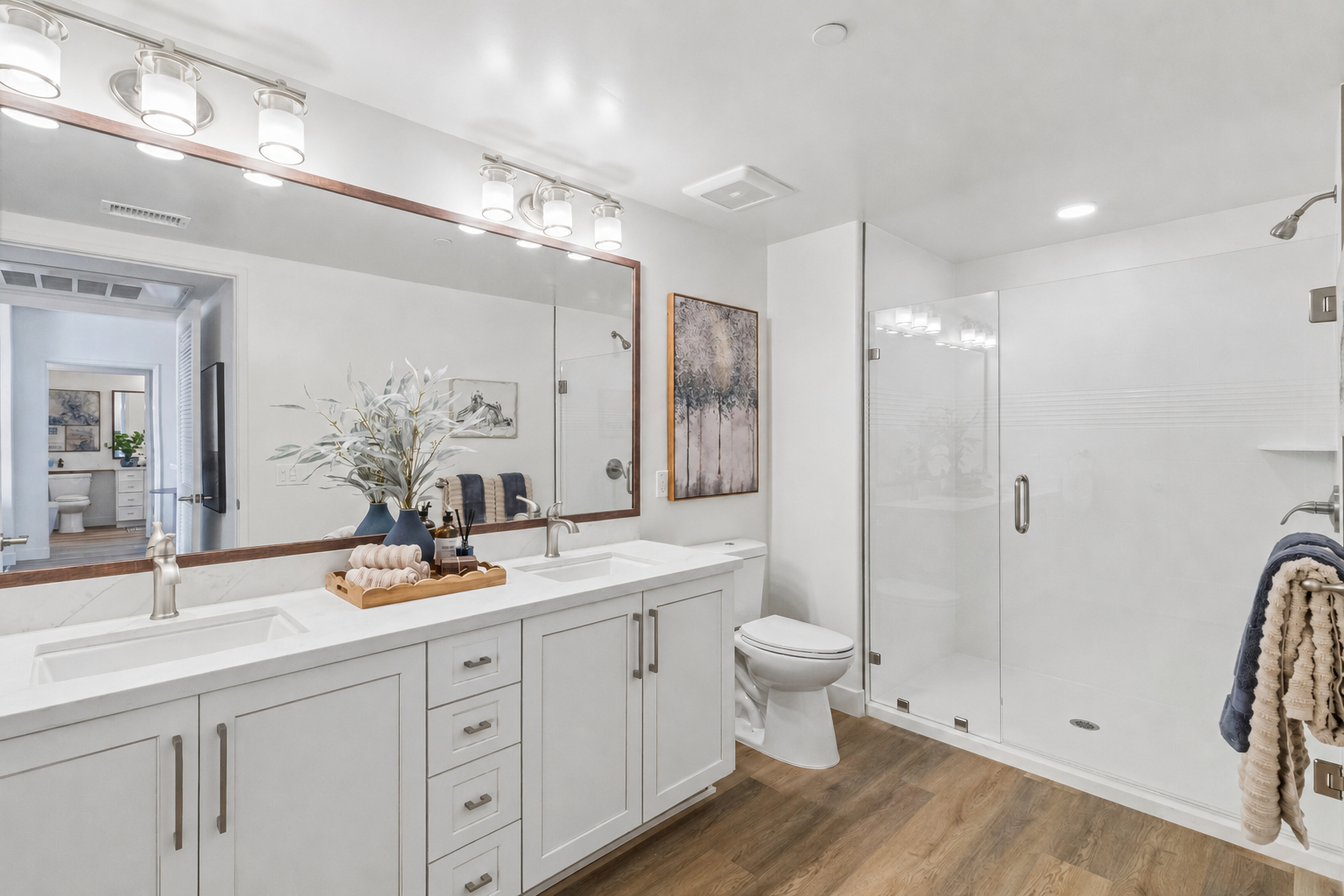 Modern bathroom featuring a dual vanity with a large mirror, stylish fixtures, and decorative elements. There is a glass shower enclosure, a toilet in a separate area, and a minimalist design with wooden flooring and soft lighting, creating a serene atmosphere.