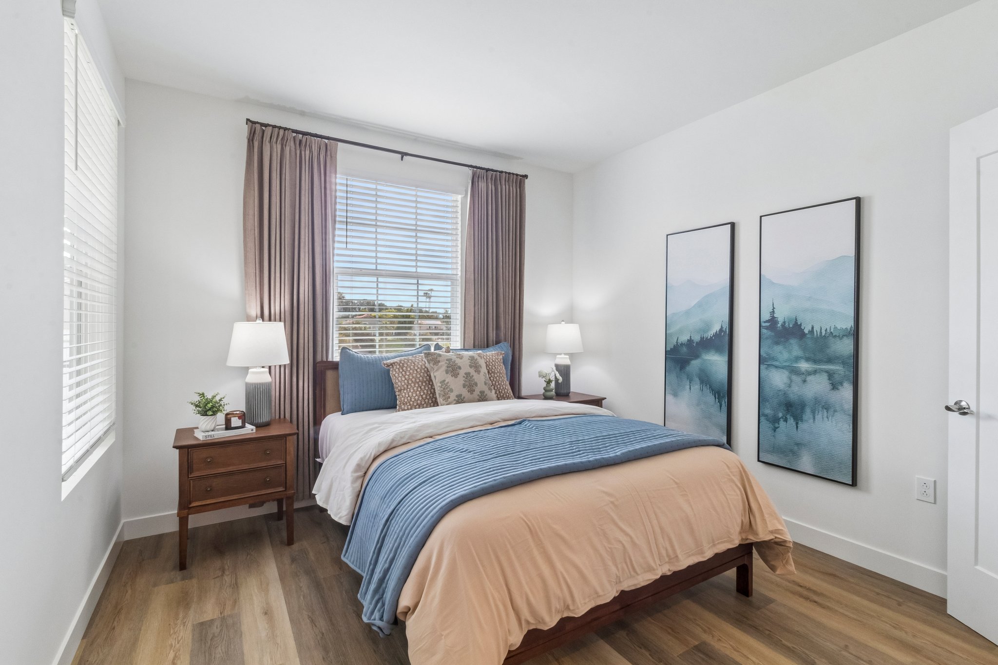 A cozy, modern bedroom featuring a neatly made bed with blue and beige bedding. There are two bedside tables with lamps on either side. Large windows with light curtains allow natural light to brighten the room. Two framed mountain landscape paintings decorate the wall, adding a serene touch to the decor.