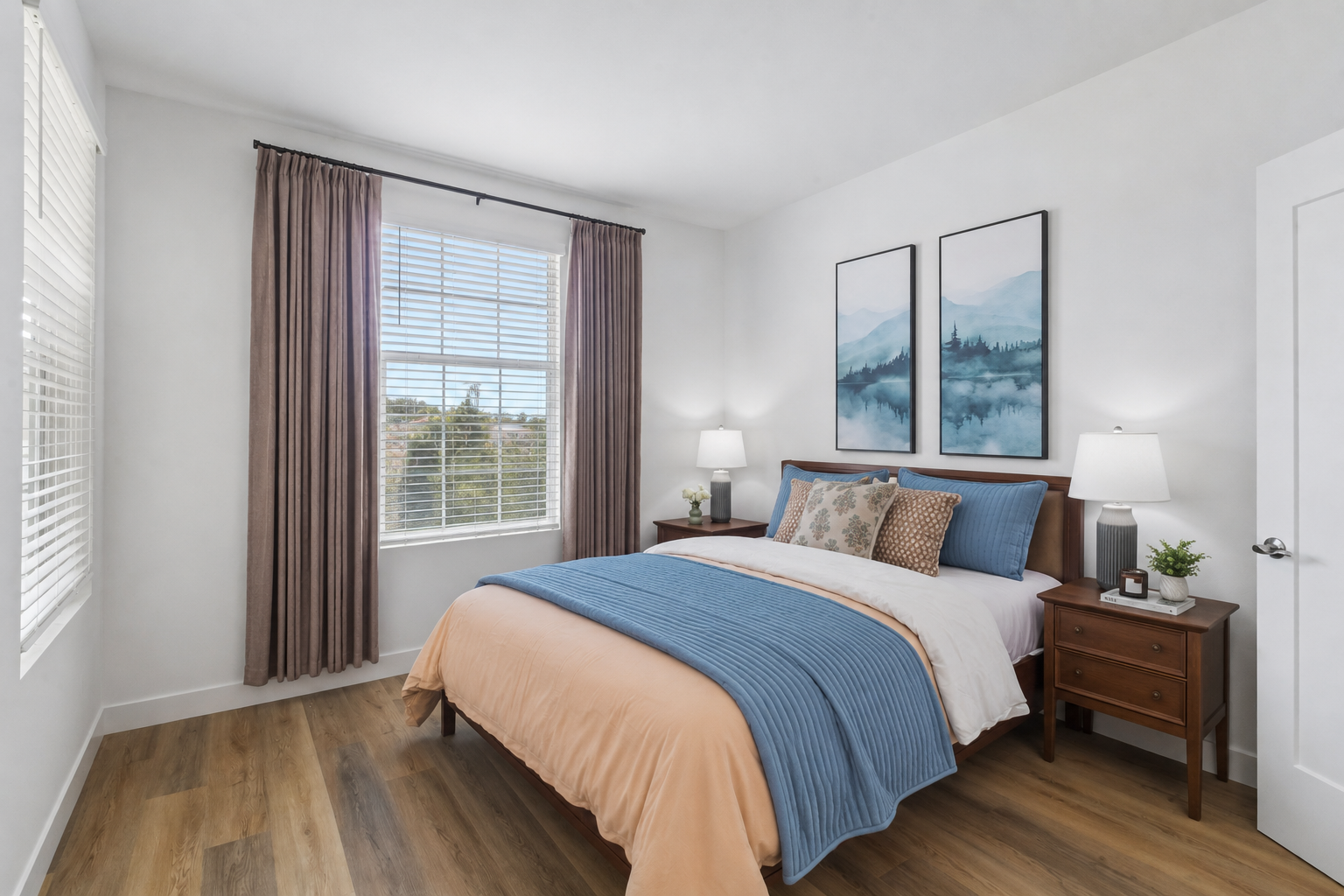 A cozy bedroom featuring a bed with blue and peach bedding, two nightstands with table lamps, and a large window with white blinds. The walls are painted light gray, and two framed mountain landscape artworks hang above the bed. Light wood flooring adds warmth to the space.
