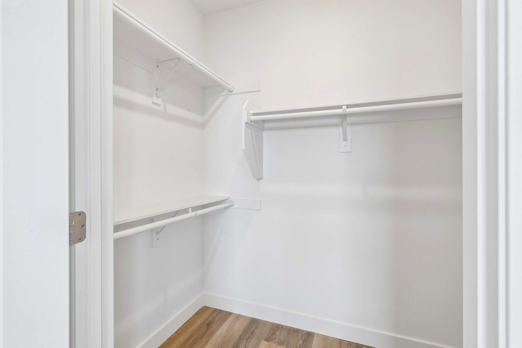 A clean, empty closet featuring white shelving and hanging rods. The walls are painted in a light color, and the floor has a wood-like finish. There are no items stored inside, creating a spacious and organized appearance.