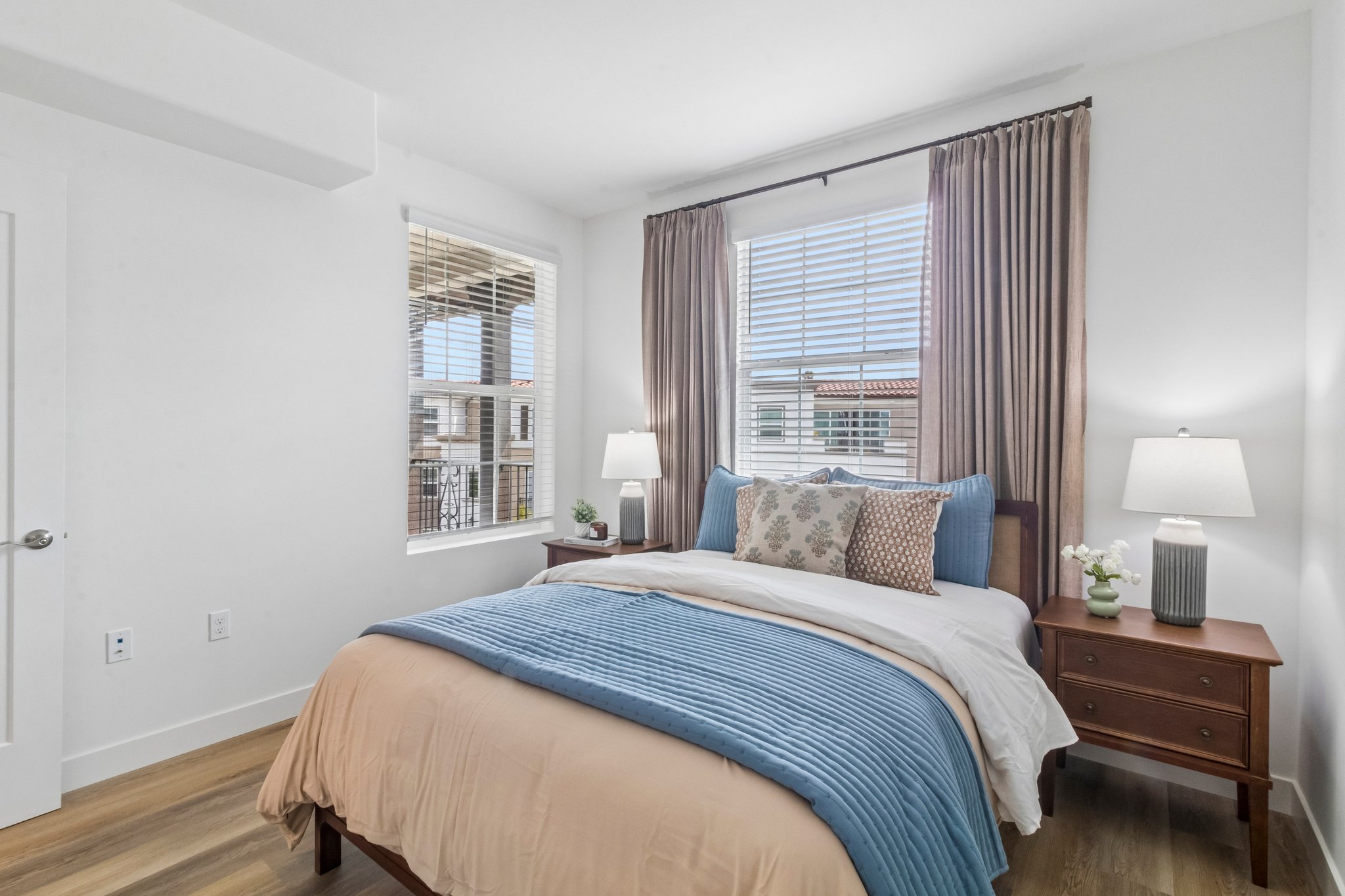 A cozy bedroom featuring a neatly made bed with blue and beige bedding, two bedside tables with lamps, and large windows with light curtains. The walls are painted white, and there are decorative pillows on the bed. Natural light brightens the room, creating a warm and inviting atmosphere.