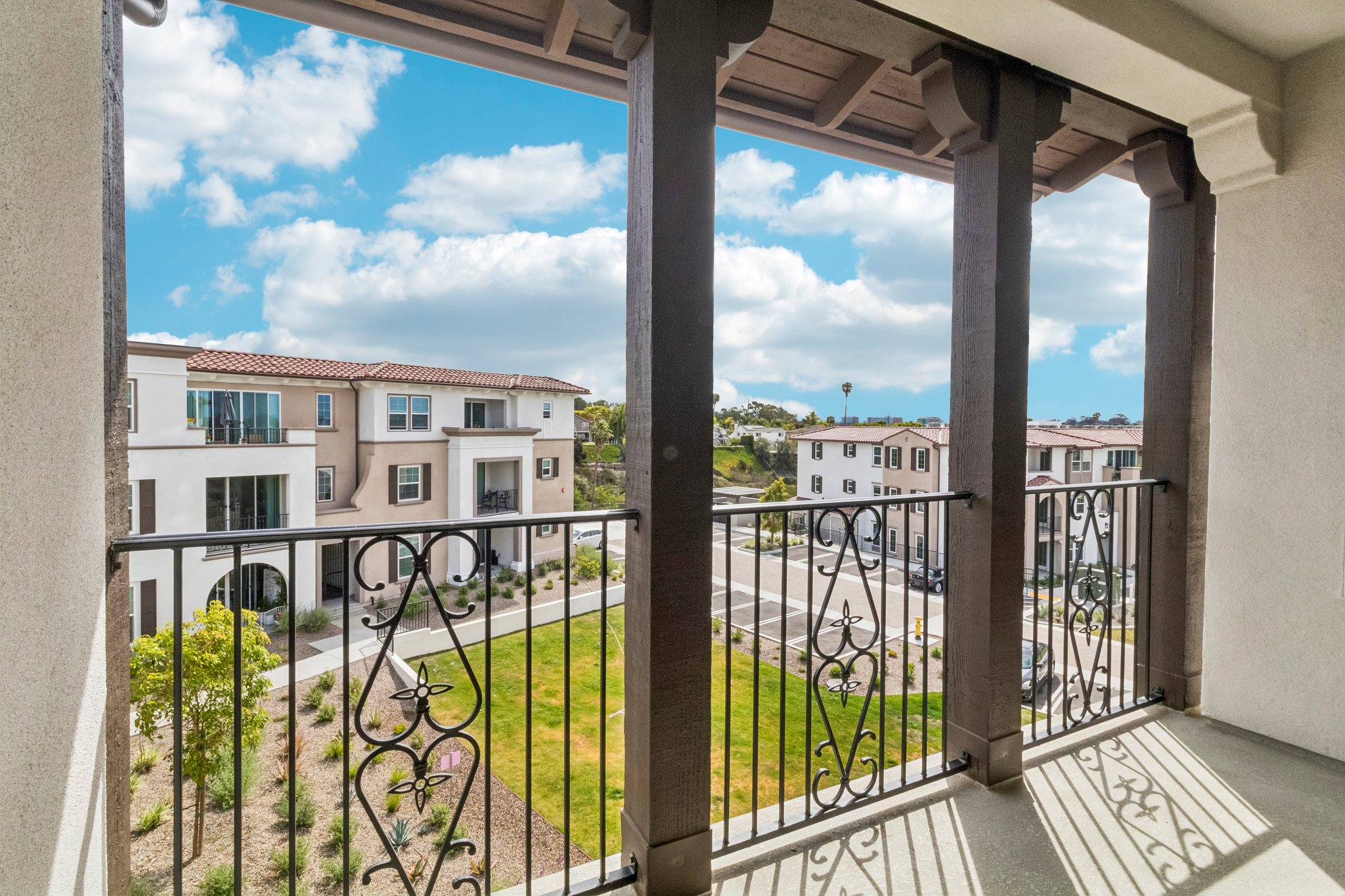 View from a balcony featuring decorative railings, overlooking a landscaped area with grass and pathways, surrounded by modern residential buildings under a partly cloudy sky.