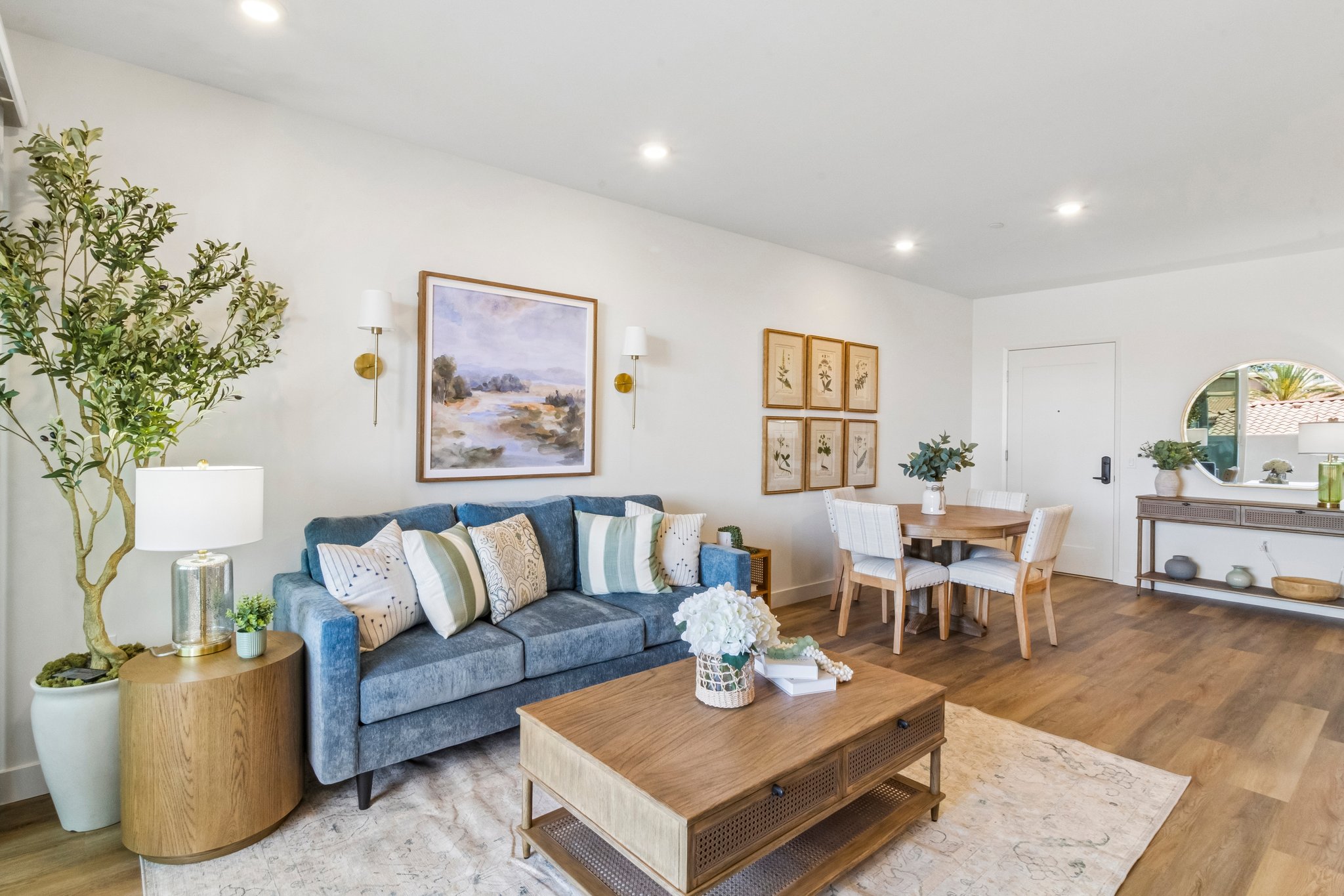 A modern living room featuring a blue sofa with decorative pillows, a wooden coffee table, and a dining area with a round table and white chairs. The room includes indoor plants, wall art with landscape paintings, and a mirror, all set against light-colored walls and warm wood flooring.