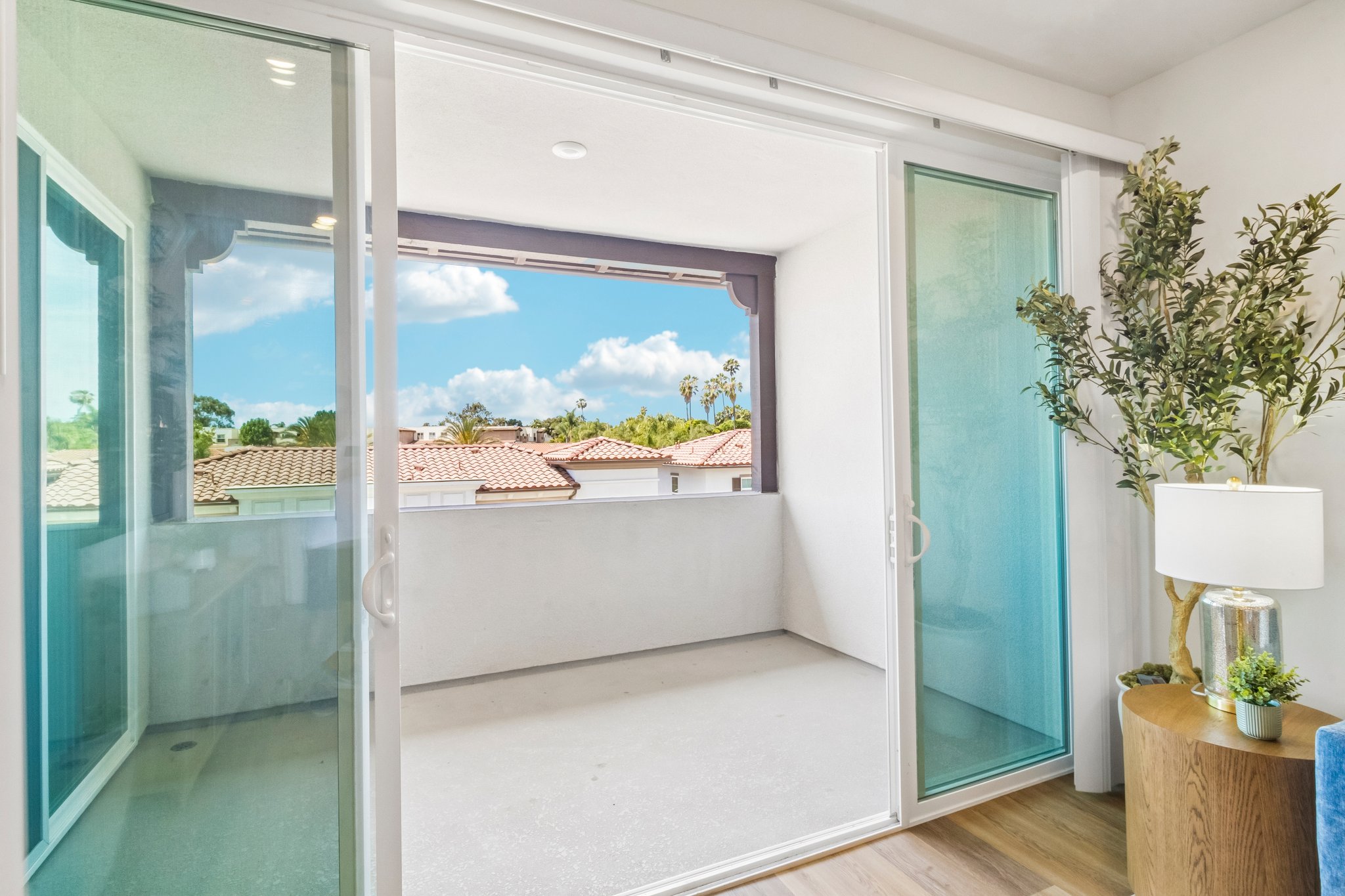 A bright and airy interior space featuring sliding glass doors that open to a small balcony. The view shows a clear sky with fluffy clouds, palm trees in the distance, and a neighborhood of houses with tiled roofs. There's a decorative plant and a lamp on a wooden side table, enhancing the inviting atmosphere.