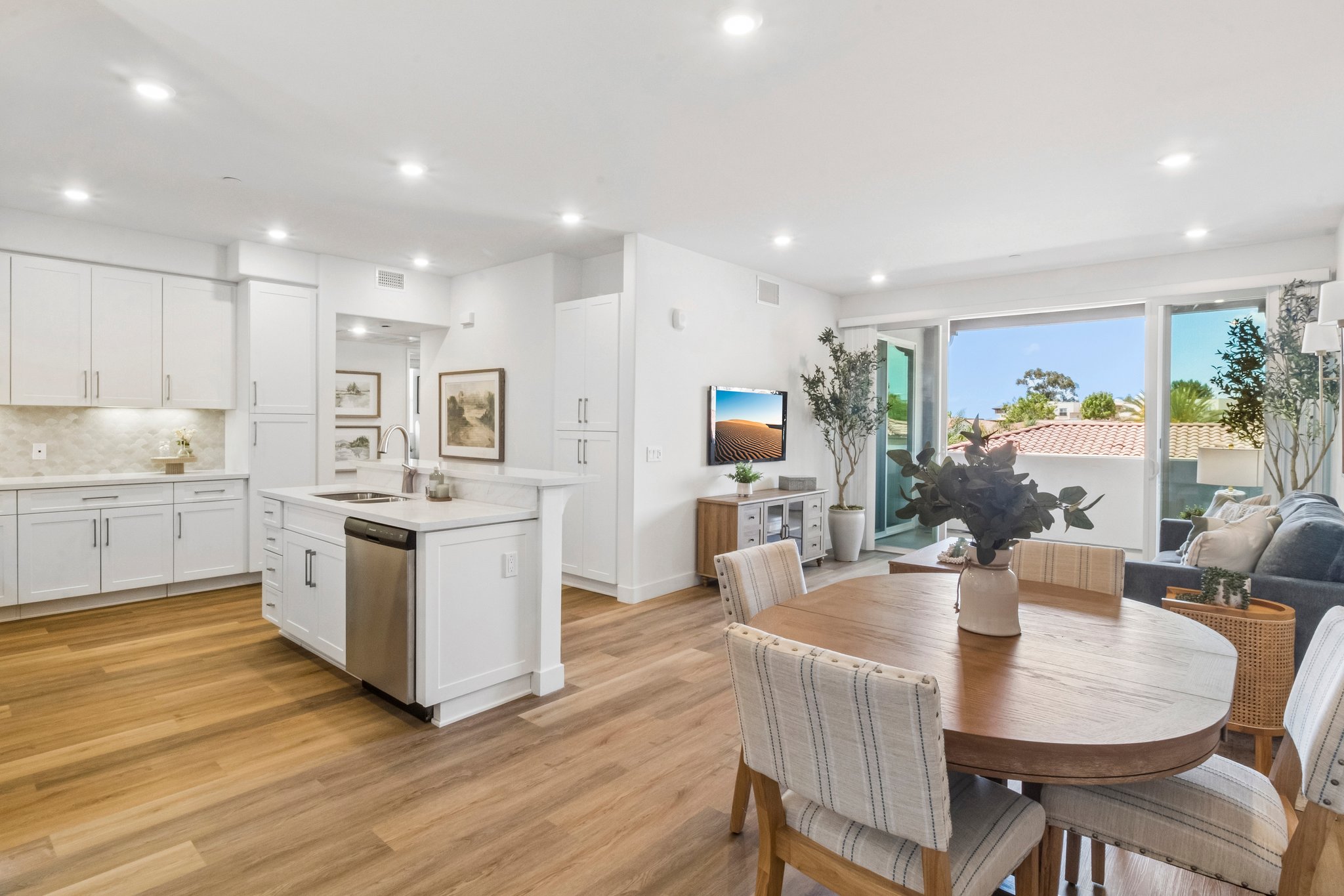 Bright and modern interior of a contemporary kitchen and living space. The kitchen features white cabinetry and stainless steel appliances. A dining table holds decorative items, and large windows allow natural light and views of greenery outside. The space has a cozy and inviting atmosphere, with stylish decor.