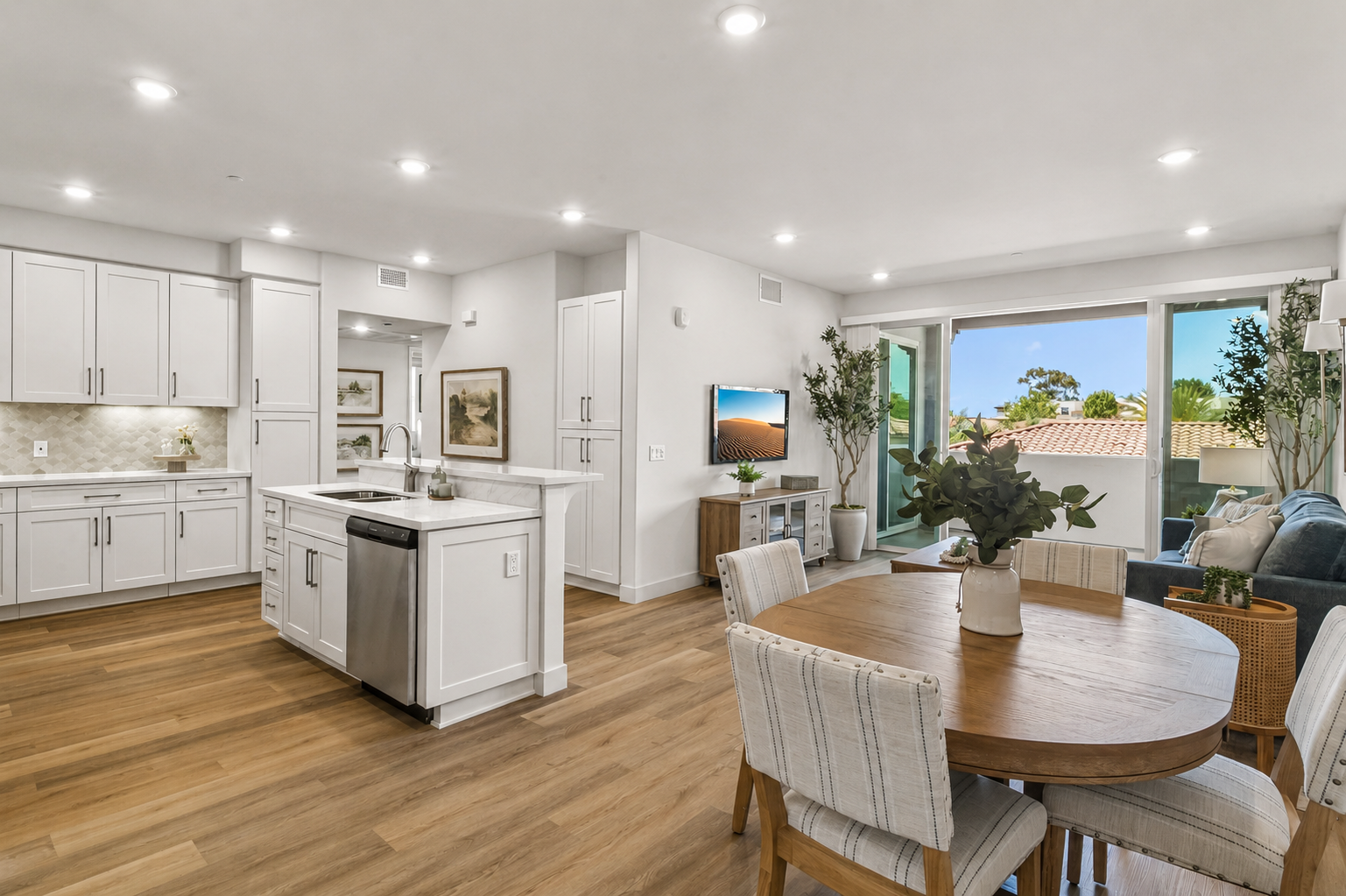 Modern kitchen and living area featuring light wood flooring, white cabinetry, and stainless steel appliances. A dining table with upholstered chairs is set in the foreground, while a cozy living space with a blue sofa and TV is visible in the background. Large windows provide natural light and a view of greenery outside.