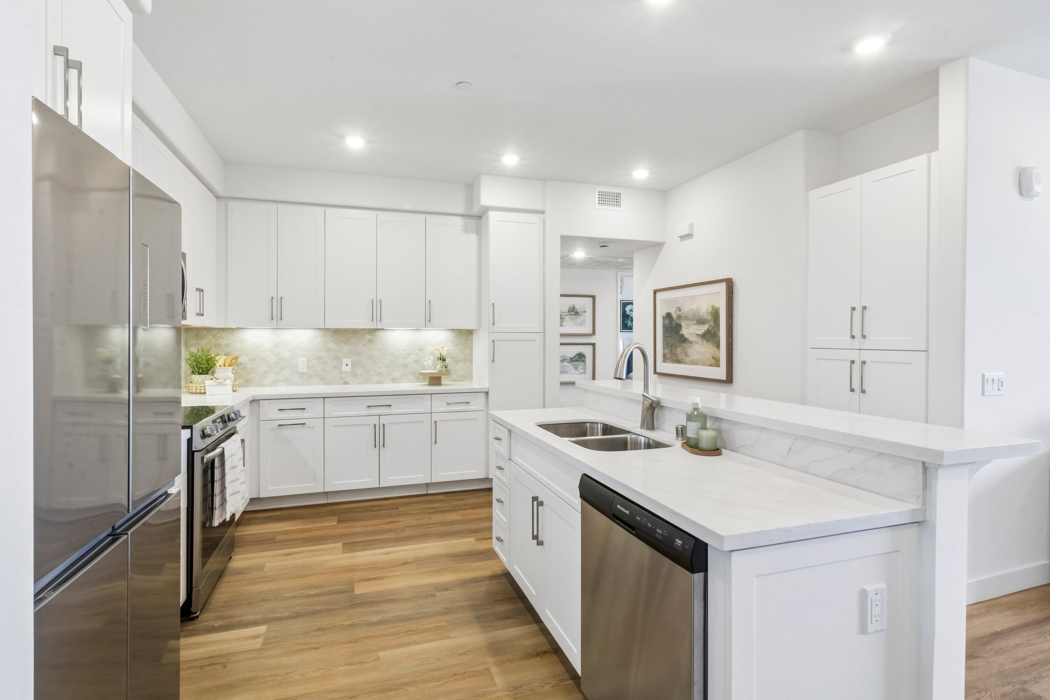 Modern kitchen featuring white cabinets, stainless steel appliances, and a large island with a marble countertop. The space is well-lit with recessed lighting, showcasing light wood flooring and decorative elements like potted plants and framed artwork on the walls.