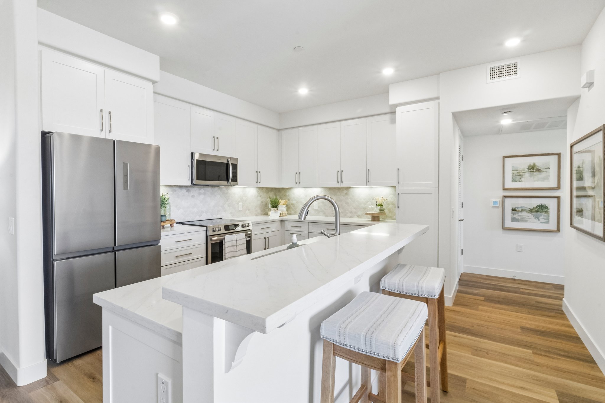 Modern kitchen featuring white cabinetry, stainless steel appliances, and a large island with bar stools. The space is well-lit with recessed lighting and has a light-colored marble countertop. The walls are painted in neutral tones, and there are framed pictures and potted plants adding decorative touches.
