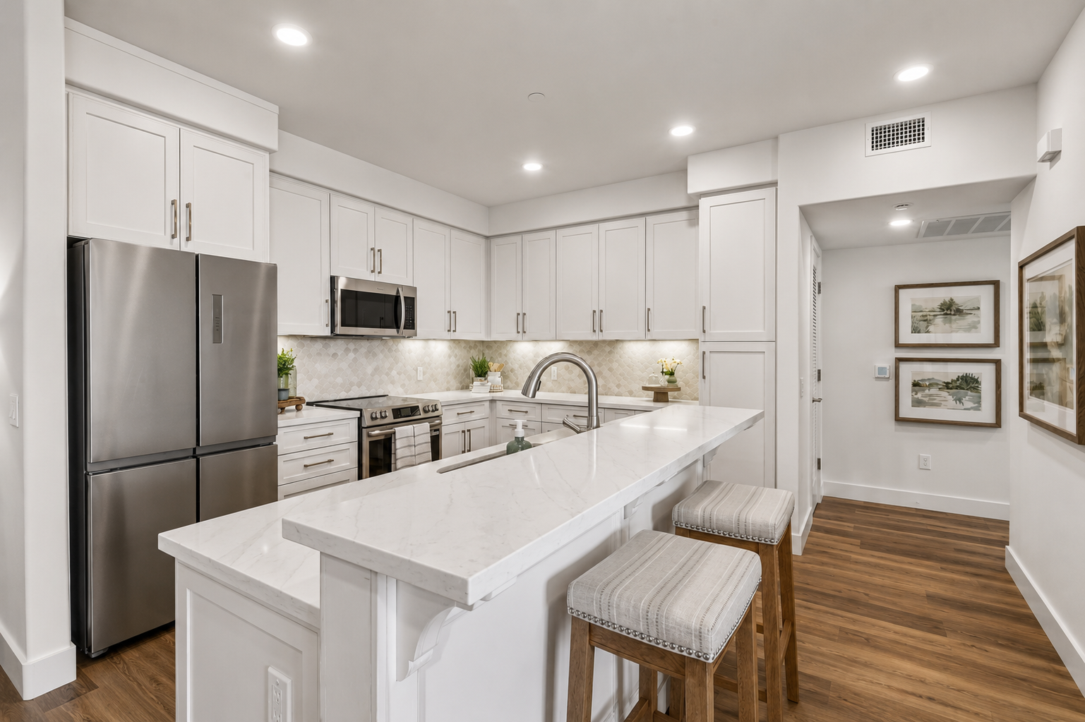 Modern kitchen featuring sleek white cabinetry, stainless steel appliances, and a large island with a marble countertop. Two barstools are positioned at the island. The space is well-lit with recessed lighting and has decorative elements like plants and framed artwork on the walls.