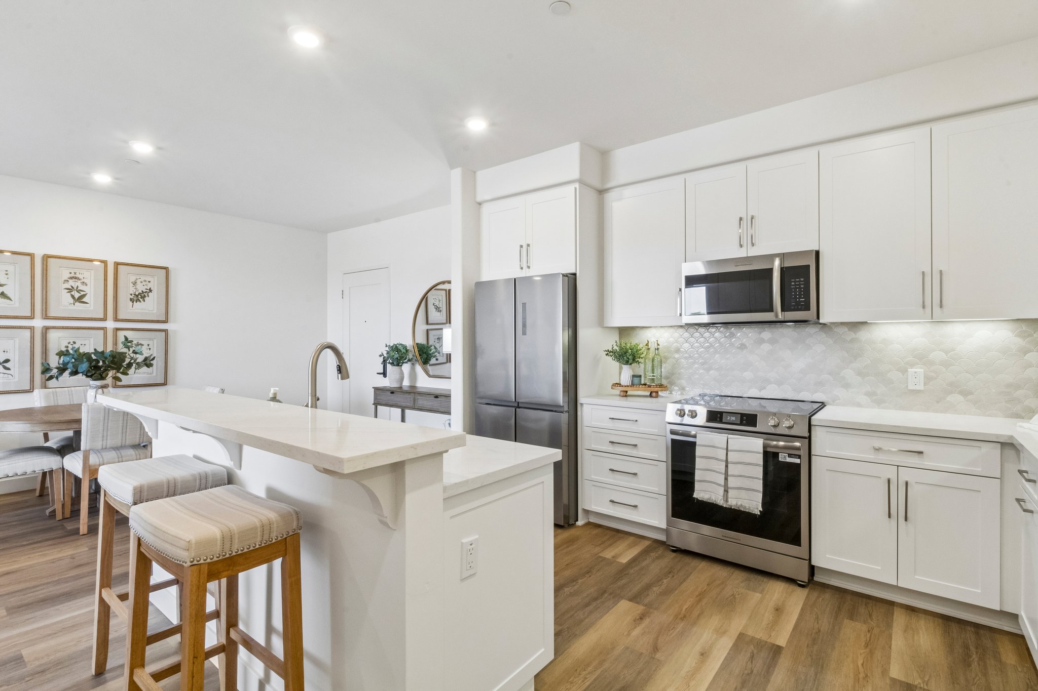 A modern kitchen featuring white cabinetry, stainless steel appliances, and a large island with bar stools. There is a dining area with a wooden table and framed botanical prints on the wall. The space is well-lit with recessed lighting and has a warm, inviting atmosphere.