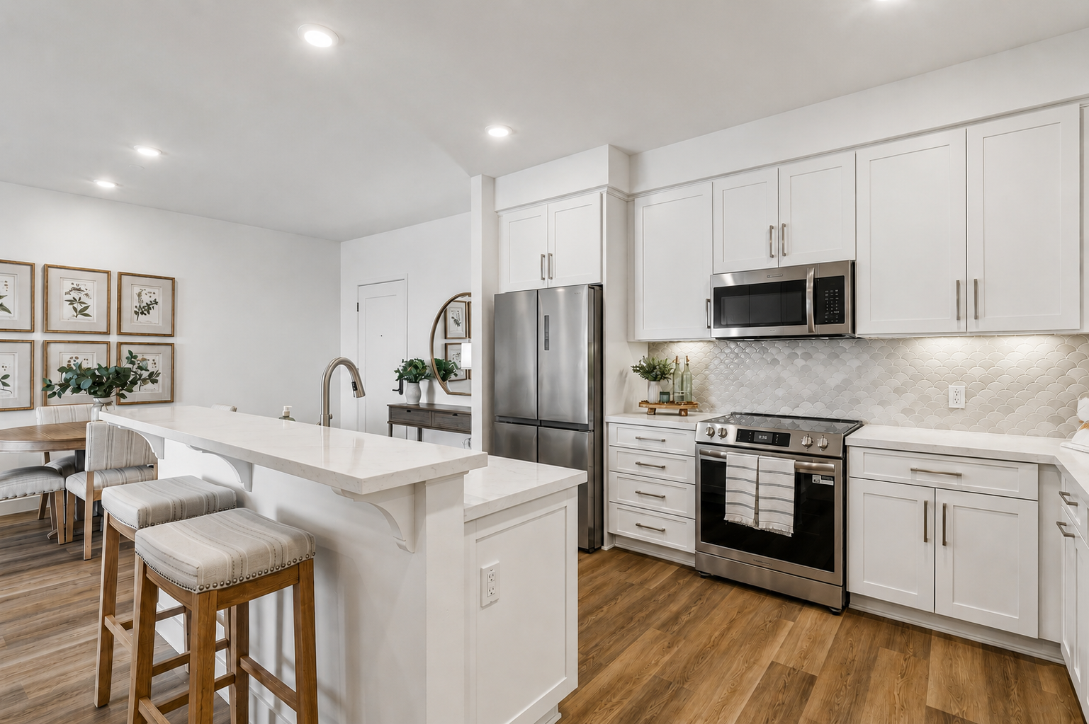 Modern kitchen featuring white cabinets, stainless steel appliances, and a quartz countertop. A central island with bar stools provides seating, while a dining area with framed artwork and greenery complements the space. Bright lighting enhances the open, airy atmosphere.
