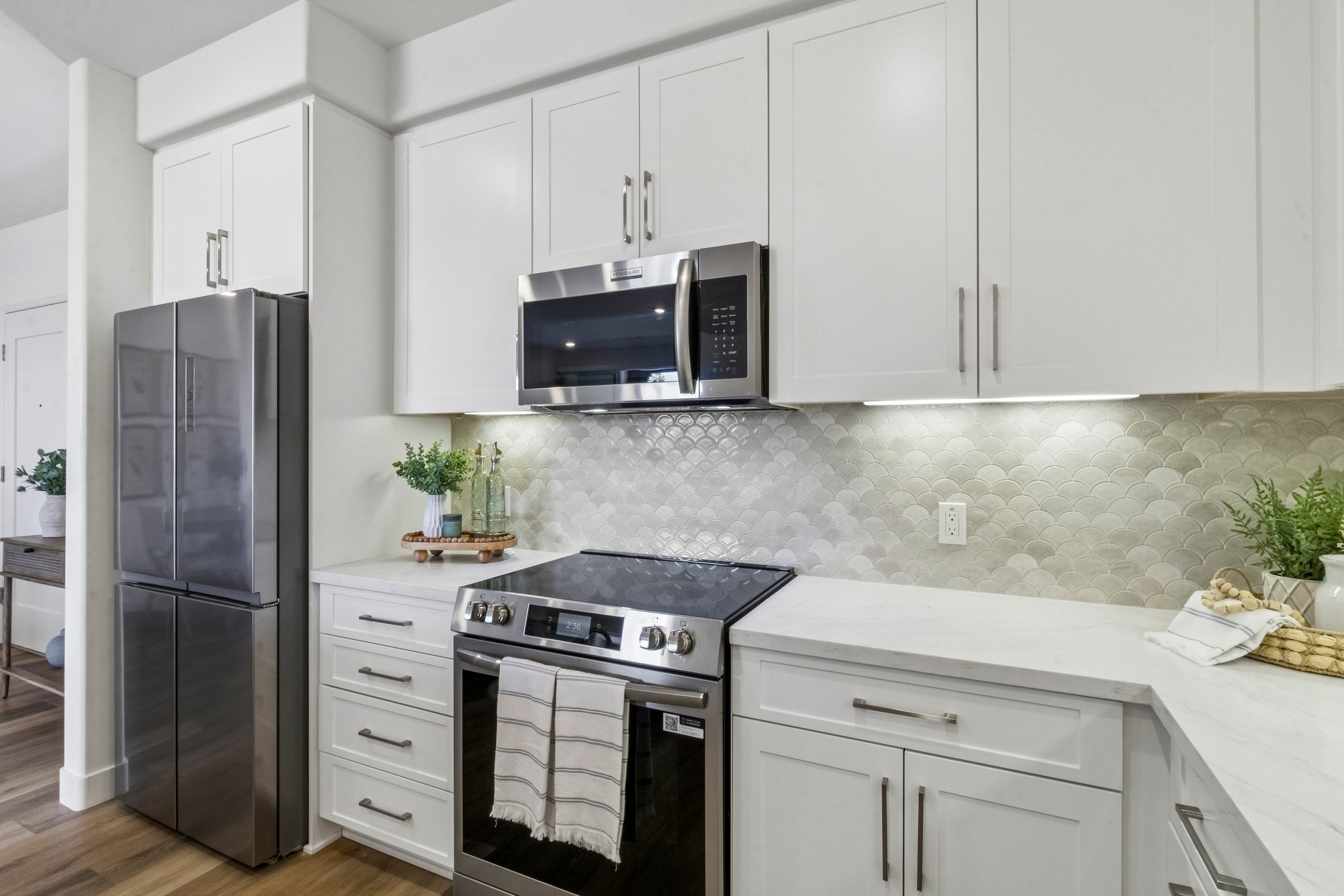 Modern kitchen featuring white cabinetry, stainless steel appliances including a refrigerator and oven, a microwave above the stove, and a stylish backsplash with scalloped tiles. The countertop is made of marble, and there are decorative plants and a towel on the counter, creating a clean and inviting atmosphere.