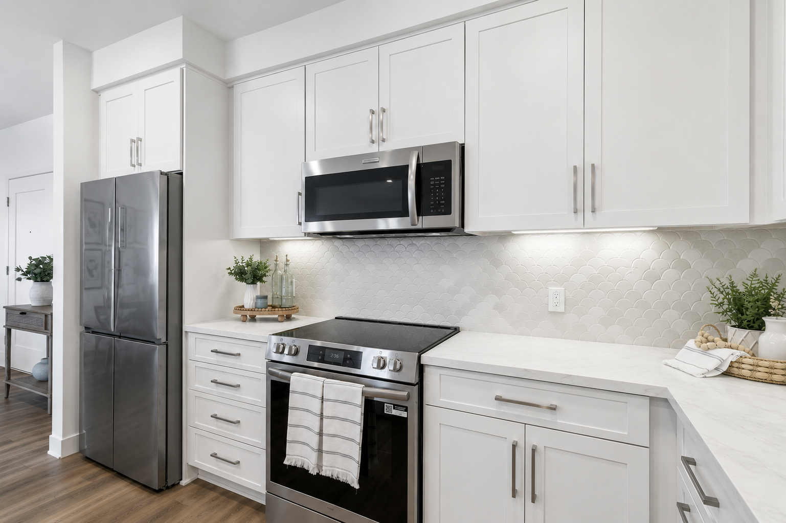 Modern kitchen featuring white cabinetry, stainless steel appliances, a black oven and stovetop, a silver refrigerator, and a microwave. The backsplash consists of light-colored, hexagonal tiles. Soft lighting enhances the clean, minimalist design, complemented by greenery in decorative pots on the countertop.