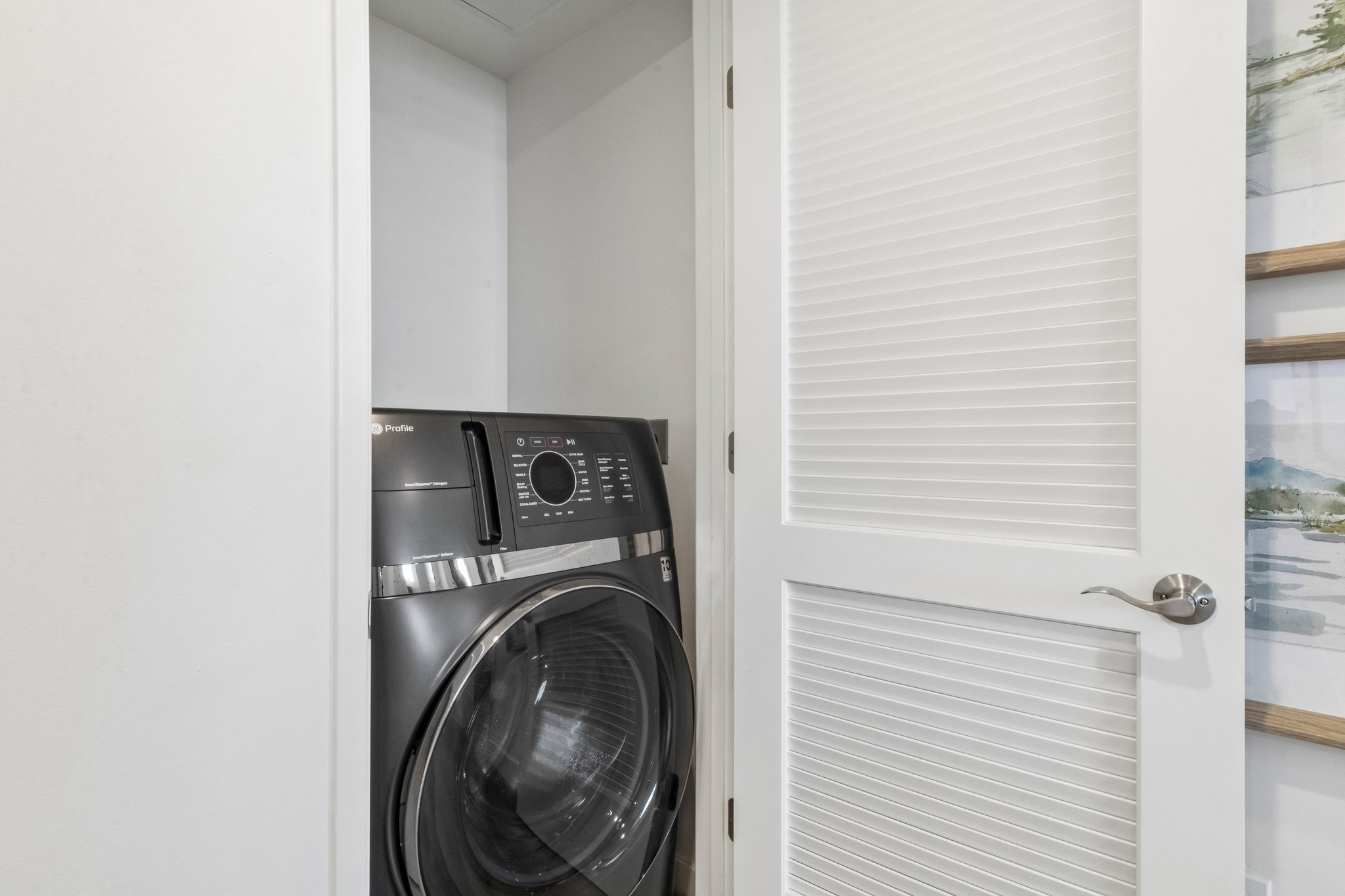 A modern black dryer inside a small laundry closet with white bi-fold doors. The interior walls are painted in a light color, and part of a wall with decorative art is visible to the right. The space is neatly organized and well-lit.