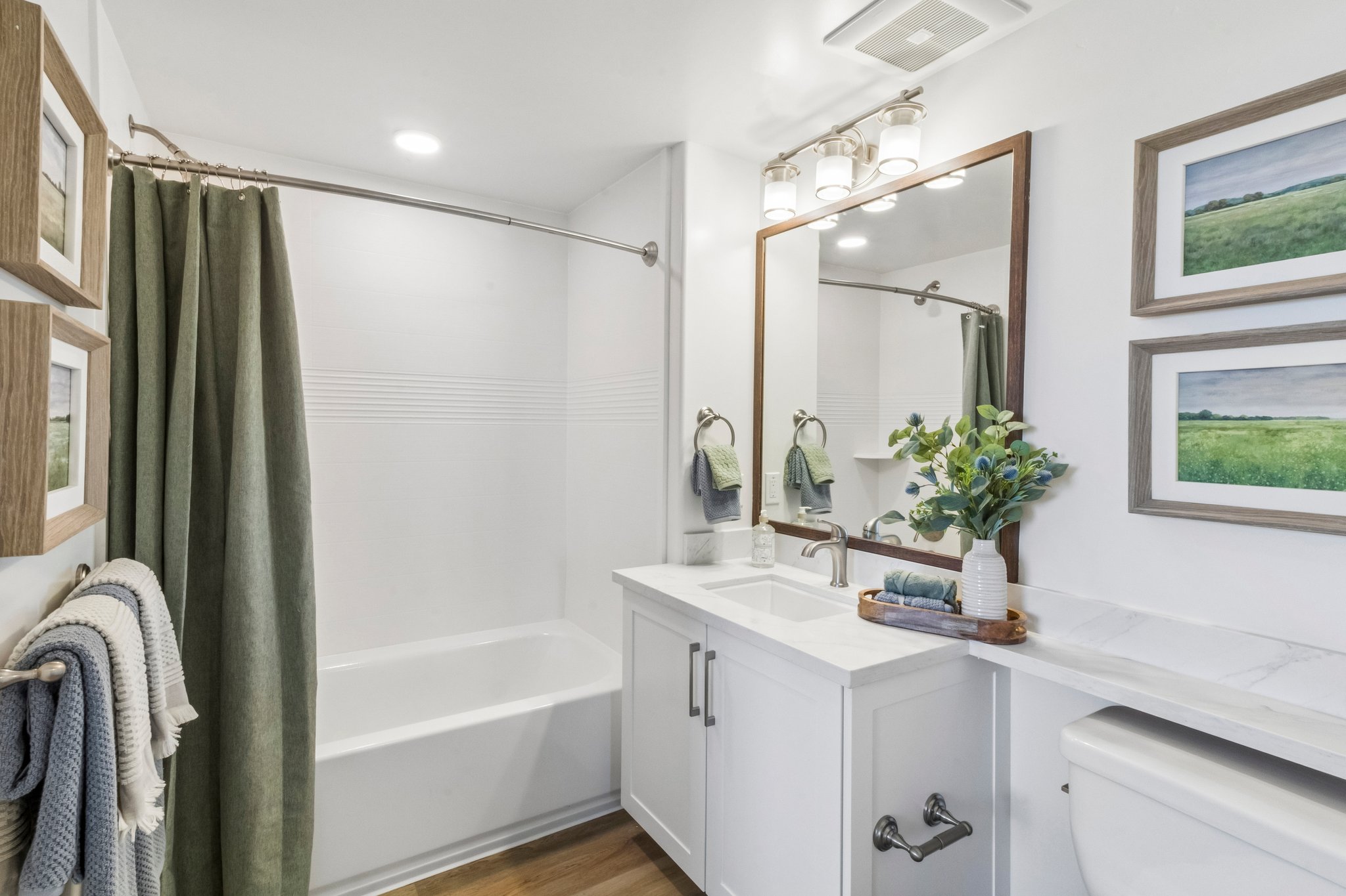 A modern bathroom featuring a white tub with a shower curtain, a double sink vanity with a marble countertop, and decorative wall art. The space is well-lit, showcasing green accents through towels and a plant, with wooden elements adding warmth. A neatly arranged towel rack is visible on the left.