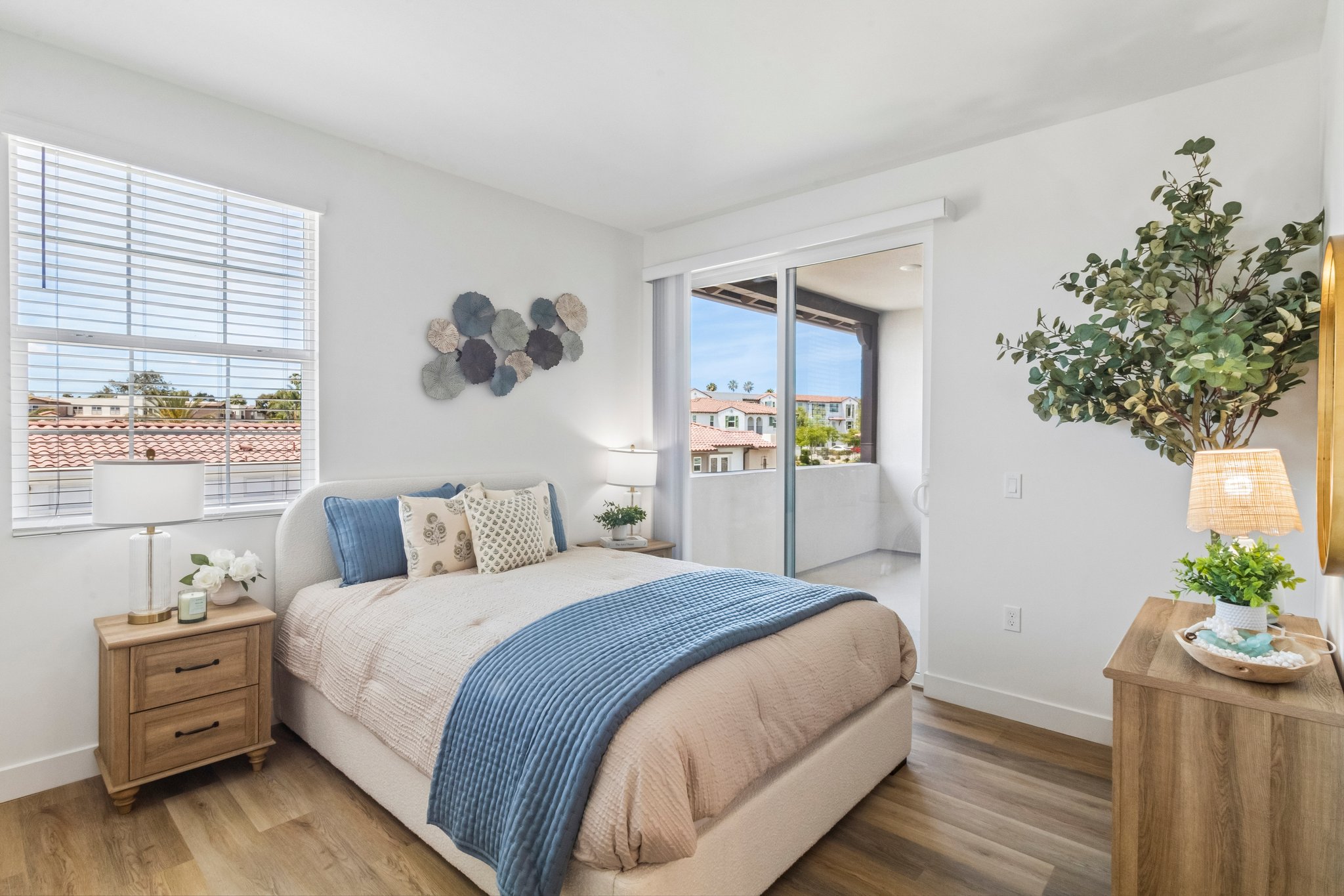 A stylish, contemporary bedroom featuring a light-colored upholstered bed with a beige and blue bedding set. There's a bedside table with a lamp, decorative plants, and artwork on the wall. A large window provides natural light, showcasing a view of buildings outside. The room has warm wood flooring and a cozy atmosphere.