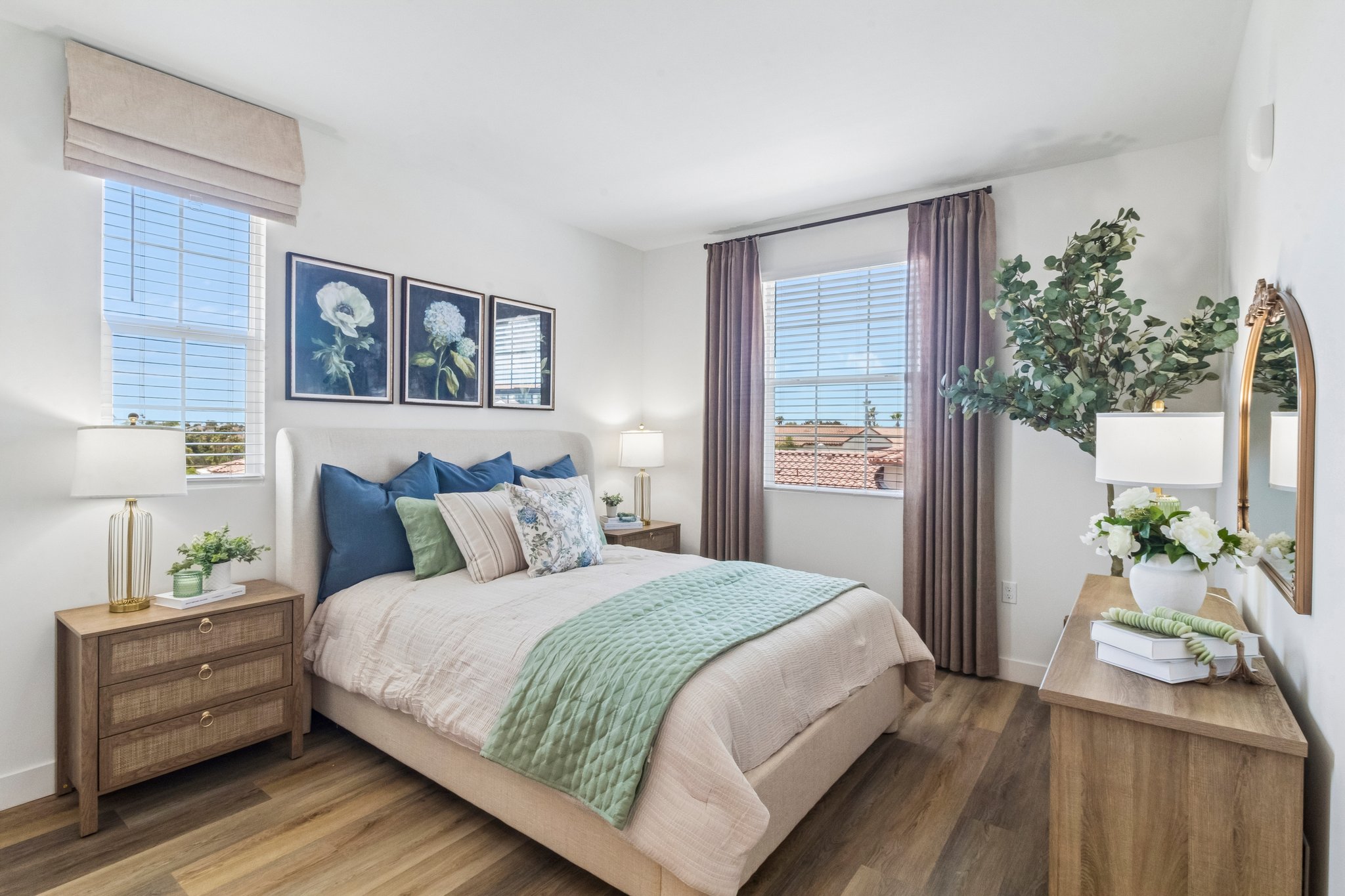 A cozy bedroom featuring a light-colored bed with blue and green accents, adorned with decorative pillows and a floral throw. There are two bedside tables with lamps, a round mirror, and framed botanical art on the wall. Large windows allow natural light to fill the room, providing views of an outdoor landscape.