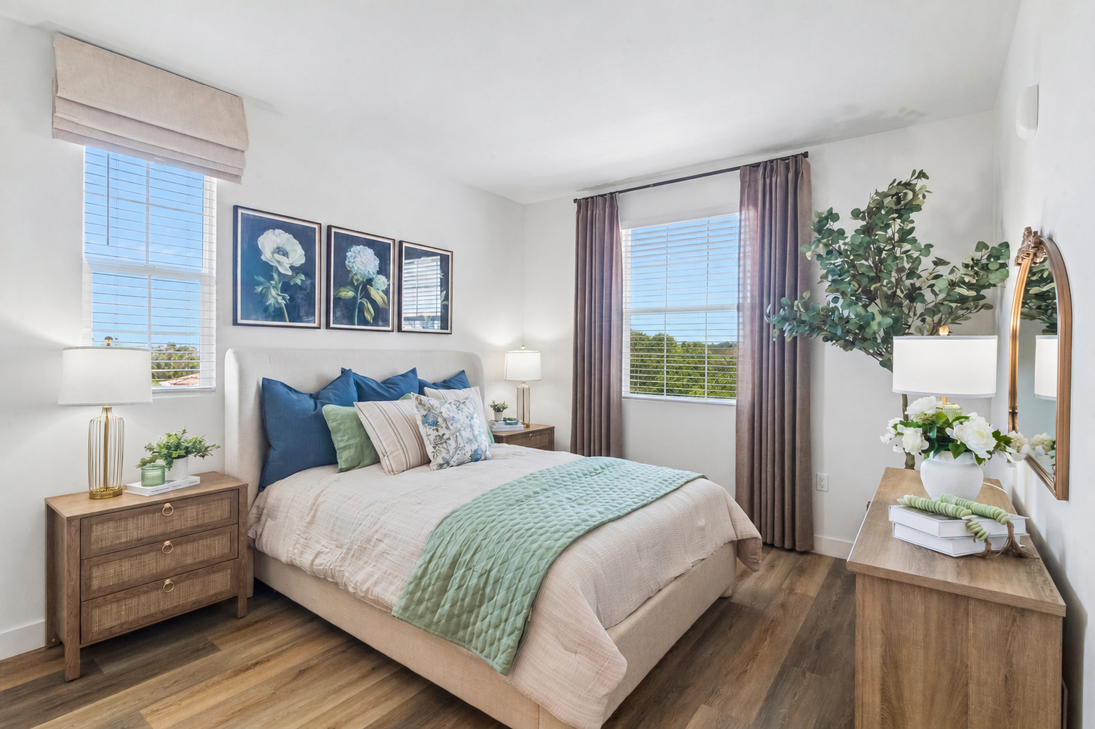 A cozy bedroom featuring a light-colored bed with decorative pillows and a quilt. Two bedside tables with lamps flank the bed, while three framed flower prints adorn the wall above. A large window with natural light showcases greenery outside, complemented by a mirror and a decorative vase on a wooden dresser.