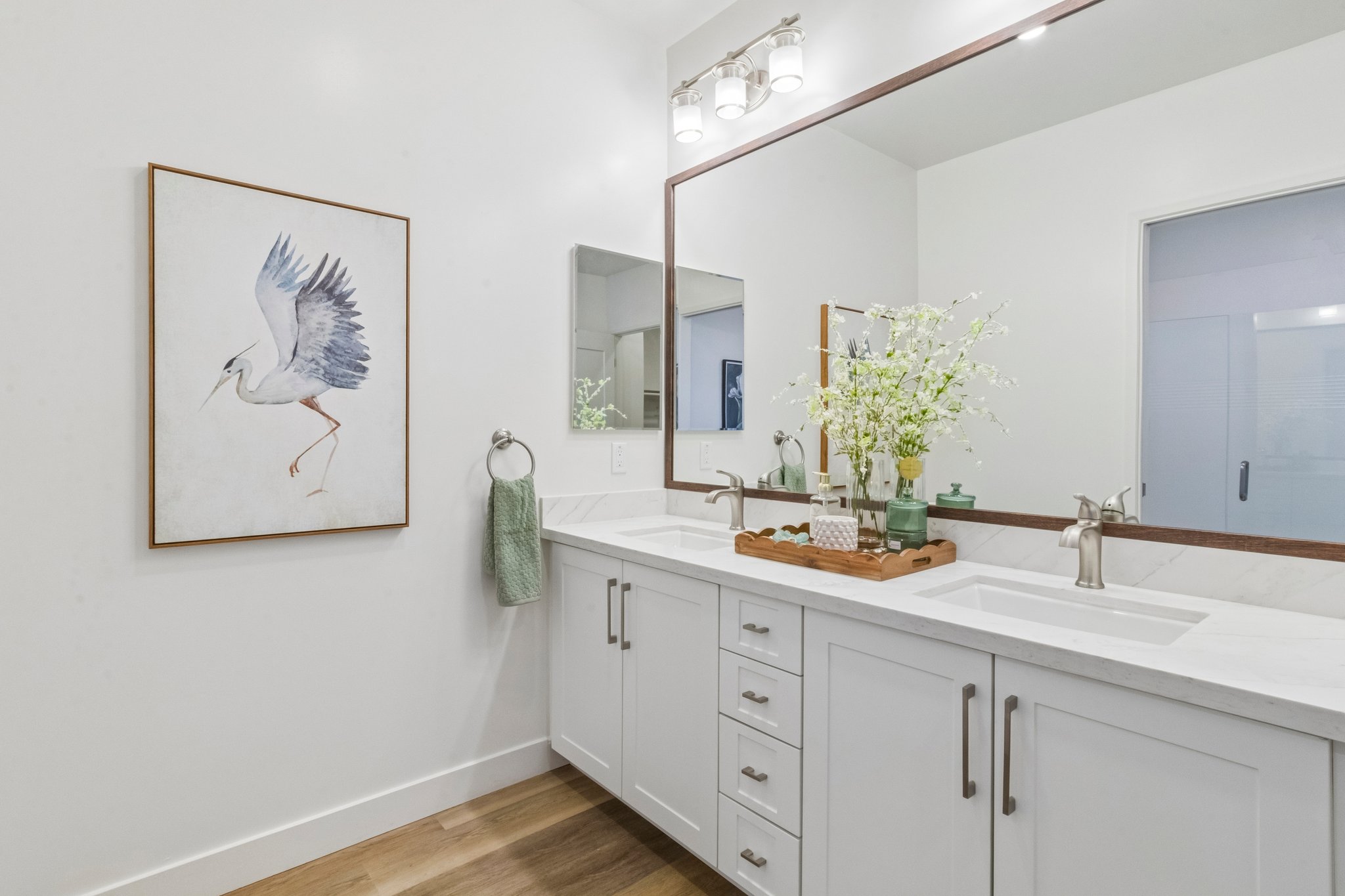 A modern bathroom featuring a white vanity with silver fixtures, a large mirror, and a decorative painting of a bird on the wall. A wooden tray holds green vases and a towel hangs on the side. The floor has light wooden planks, and there's a frosted glass door to another room.