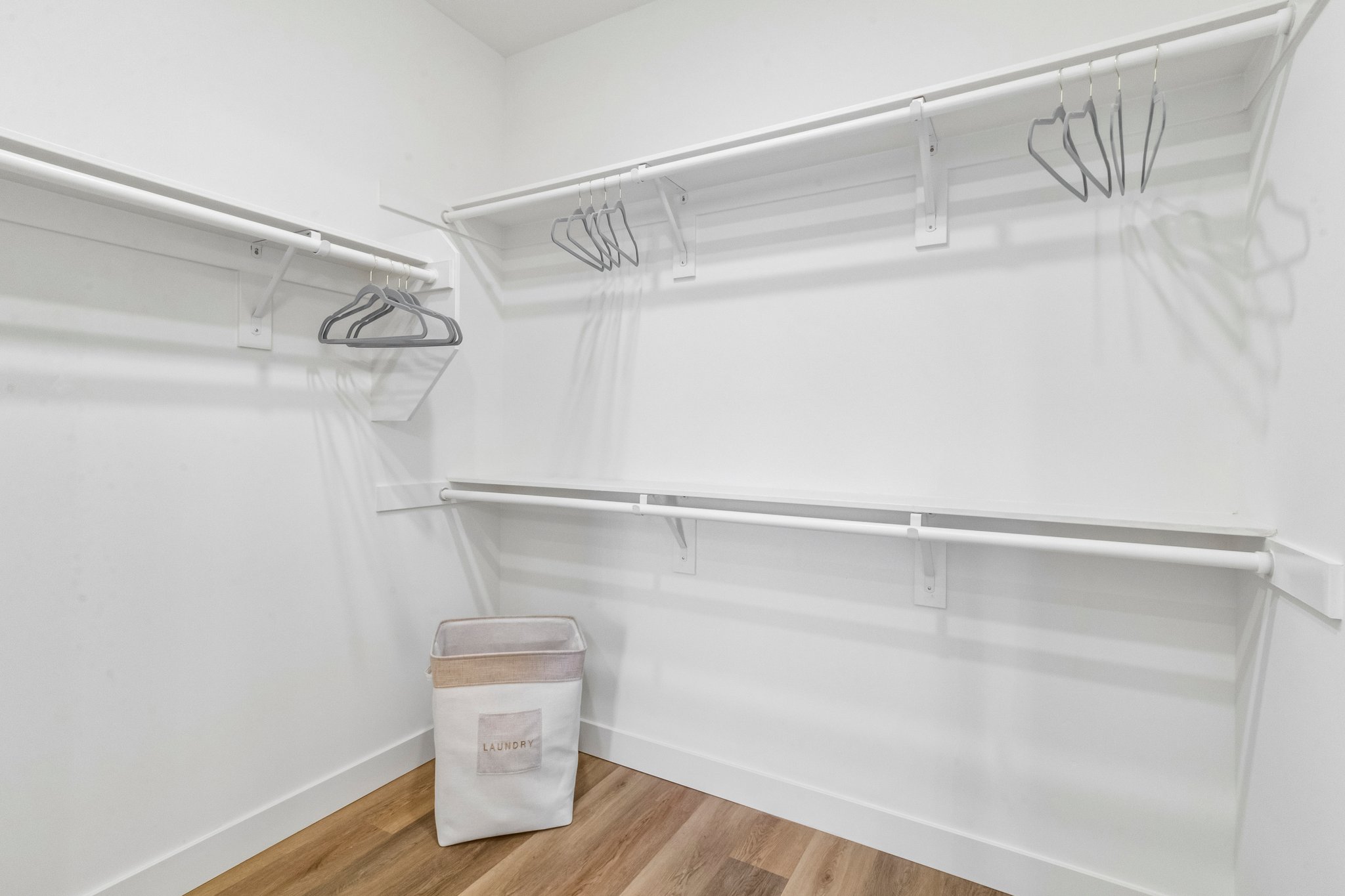An empty closet featuring white shelving and hanging rods. A few hangers are mounted on the rods, and a fabric storage bin labeled "LAUNDRY" sits on the floor, all against a backdrop of light-colored walls and wood-like flooring. The space is bright and minimalistic.