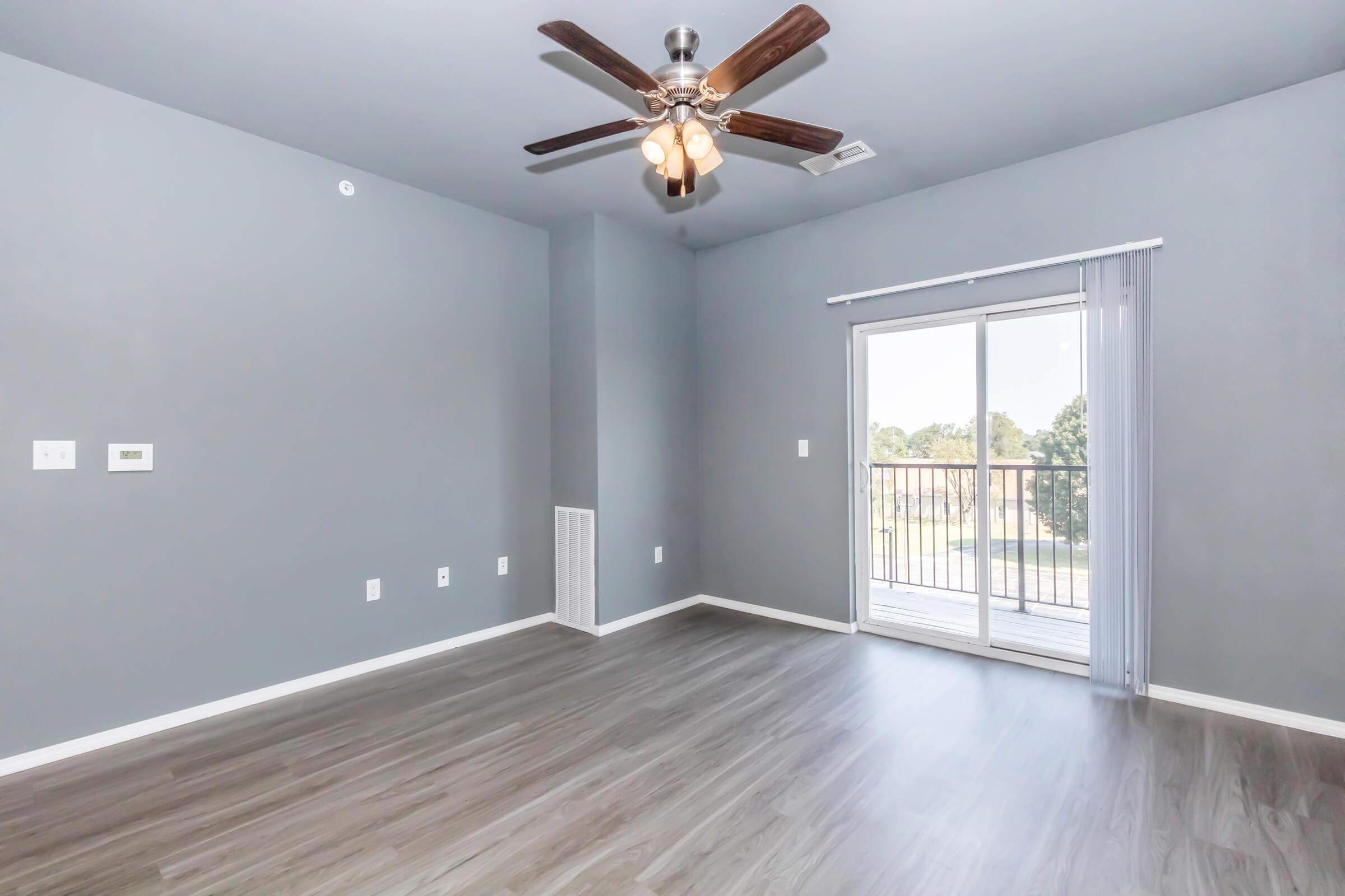A spacious, empty room with gray walls and a ceiling fan. It features a sliding glass door leading to a balcony, and modern wood-like flooring. There are electrical outlets on the wall and a vent for heating or cooling. The room is well-lit with natural light coming through the sliding door.