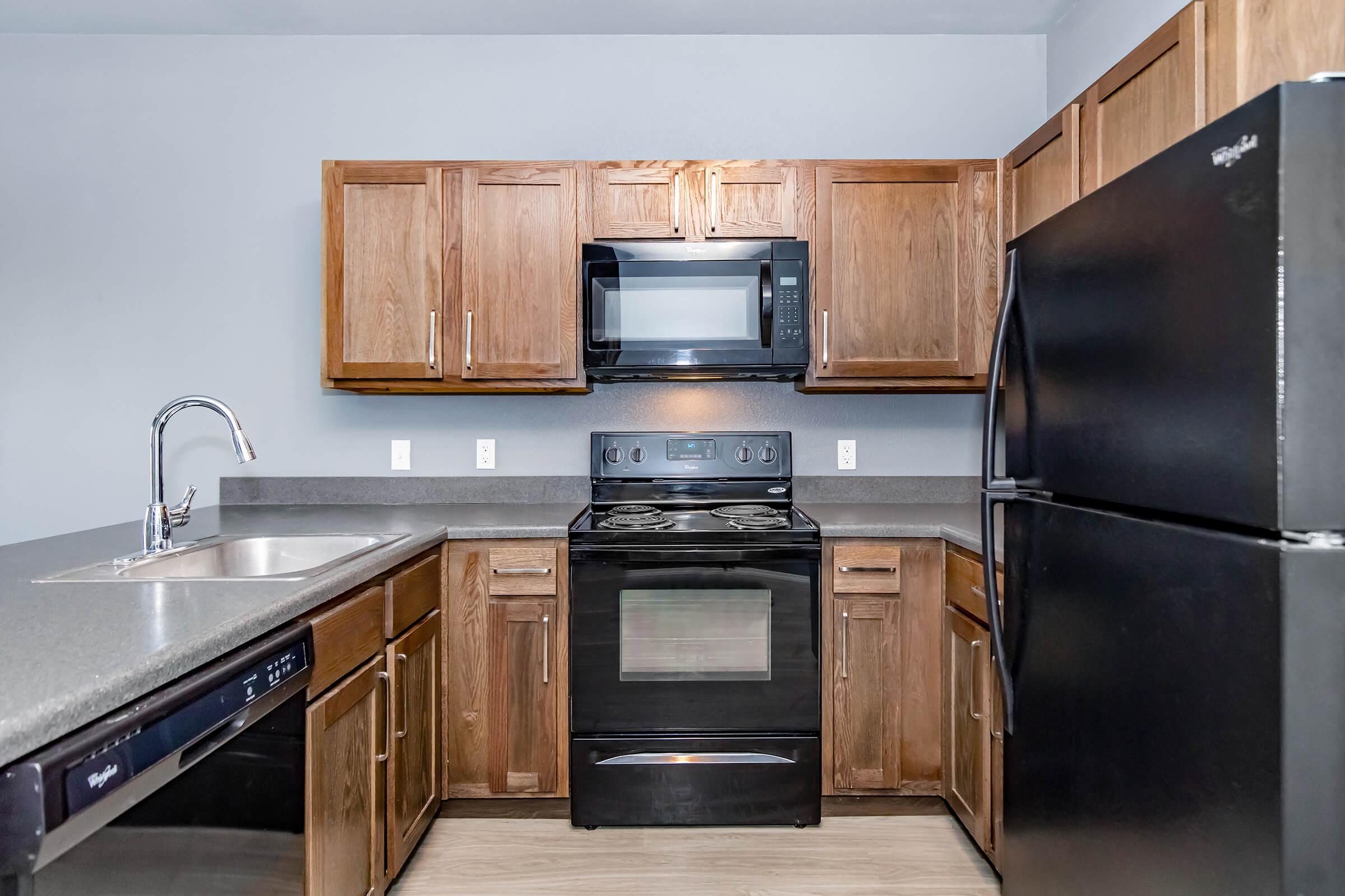 Modern kitchen featuring wooden cabinets, a black stove, microwave, and refrigerator. The countertop is dark, and there's a stainless steel sink with a faucet. The kitchen has a minimalist design with gray walls and a clean, organized layout.