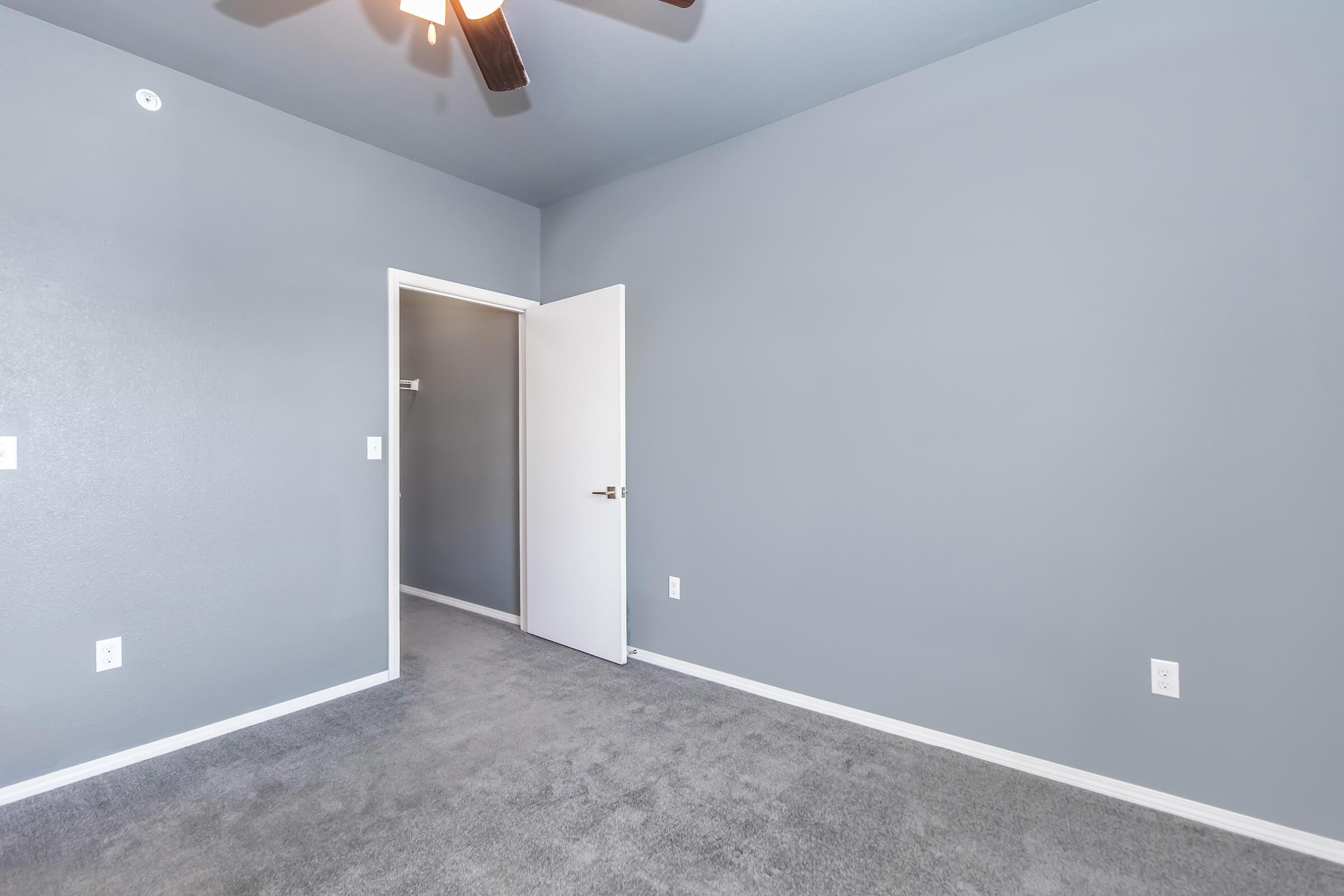 A vacant room with light gray walls and gray carpeting. The space features an open door leading to another room, a ceiling fan, and minimal furnishings. The overall appearance is clean and uncluttered, suggesting a modern, simple design suitable for various uses.
