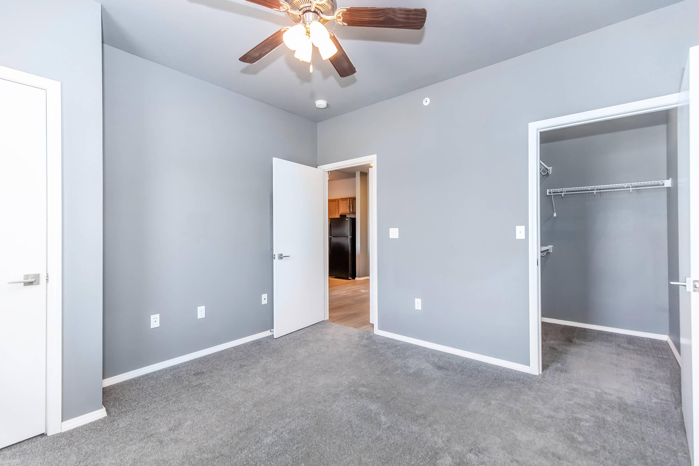 A spacious room with gray walls and light gray carpet. The ceiling features a ceiling fan, and there are two doors: one leading to a closet and another to an adjoining area. Natural light enters from the open doorway, which reveals a glimpse of a kitchen.
