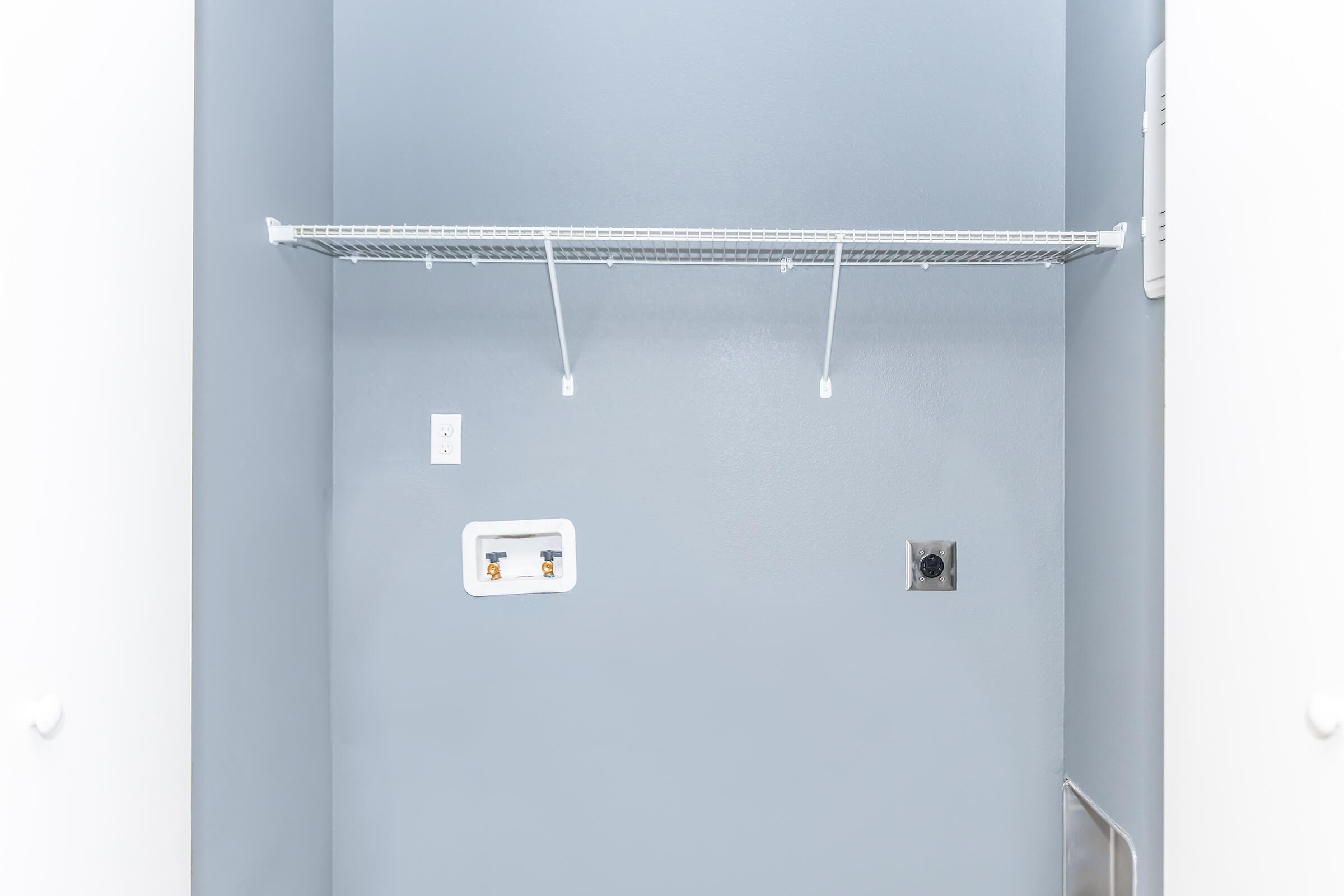 An empty laundry room with blue walls. There is a wire shelf mounted on the wall, and two brackets hanging from the shelf. A power outlet and a utility hookup are visible along the wall. The floor is bare, and no other items or appliances are present in the space.