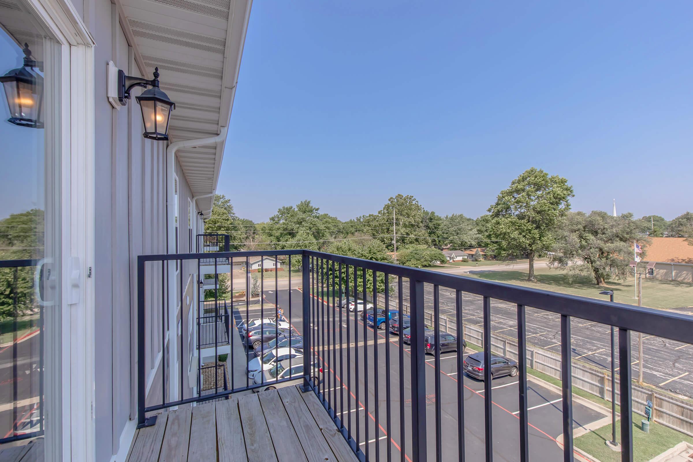 View from a balcony overlooking a parking lot and green trees under a clear blue sky. The balcony features black railing and outdoor lights. Nearby buildings and a well-maintained landscape are visible, creating a peaceful setting.