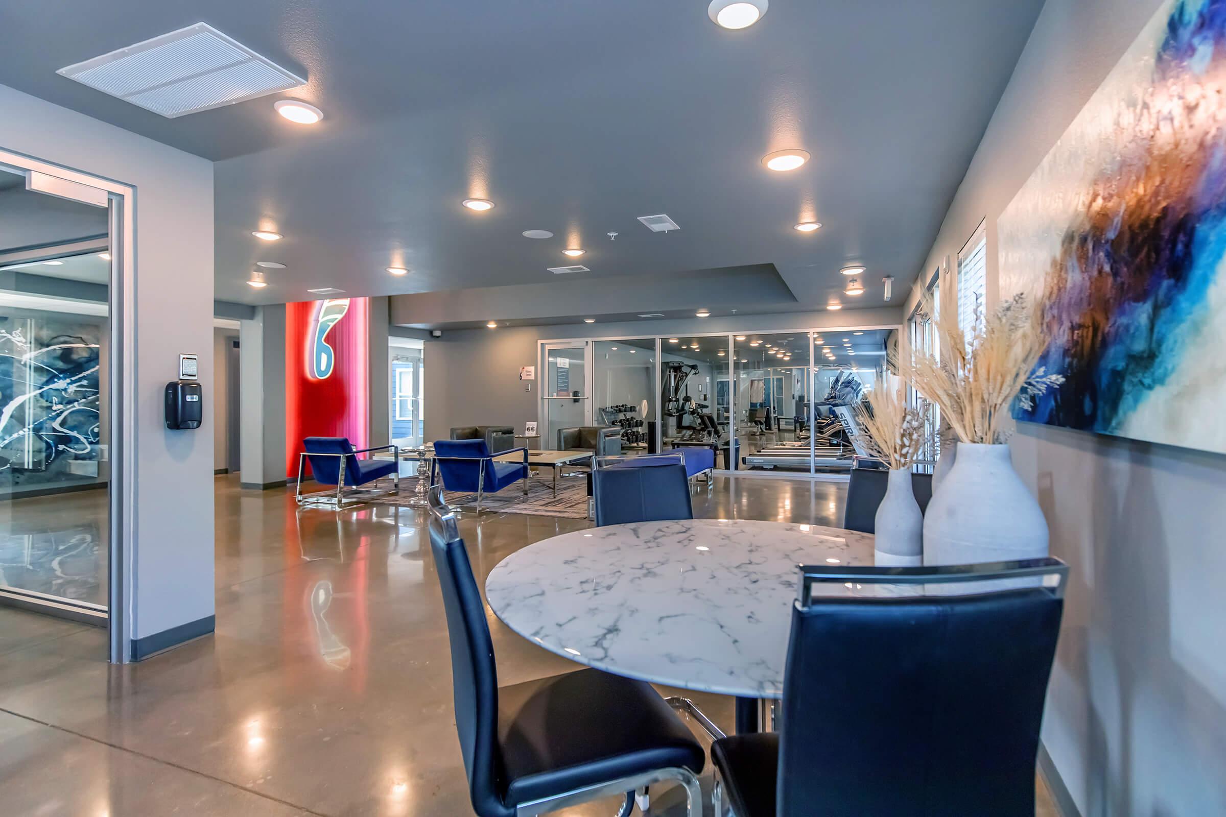 A modern indoor space featuring a round marble table with black chairs, surrounded by stylish decor. On one side, there are contemporary exercise equipment and large windows. The walls display abstract art, and soft lighting enhances the sleek atmosphere.