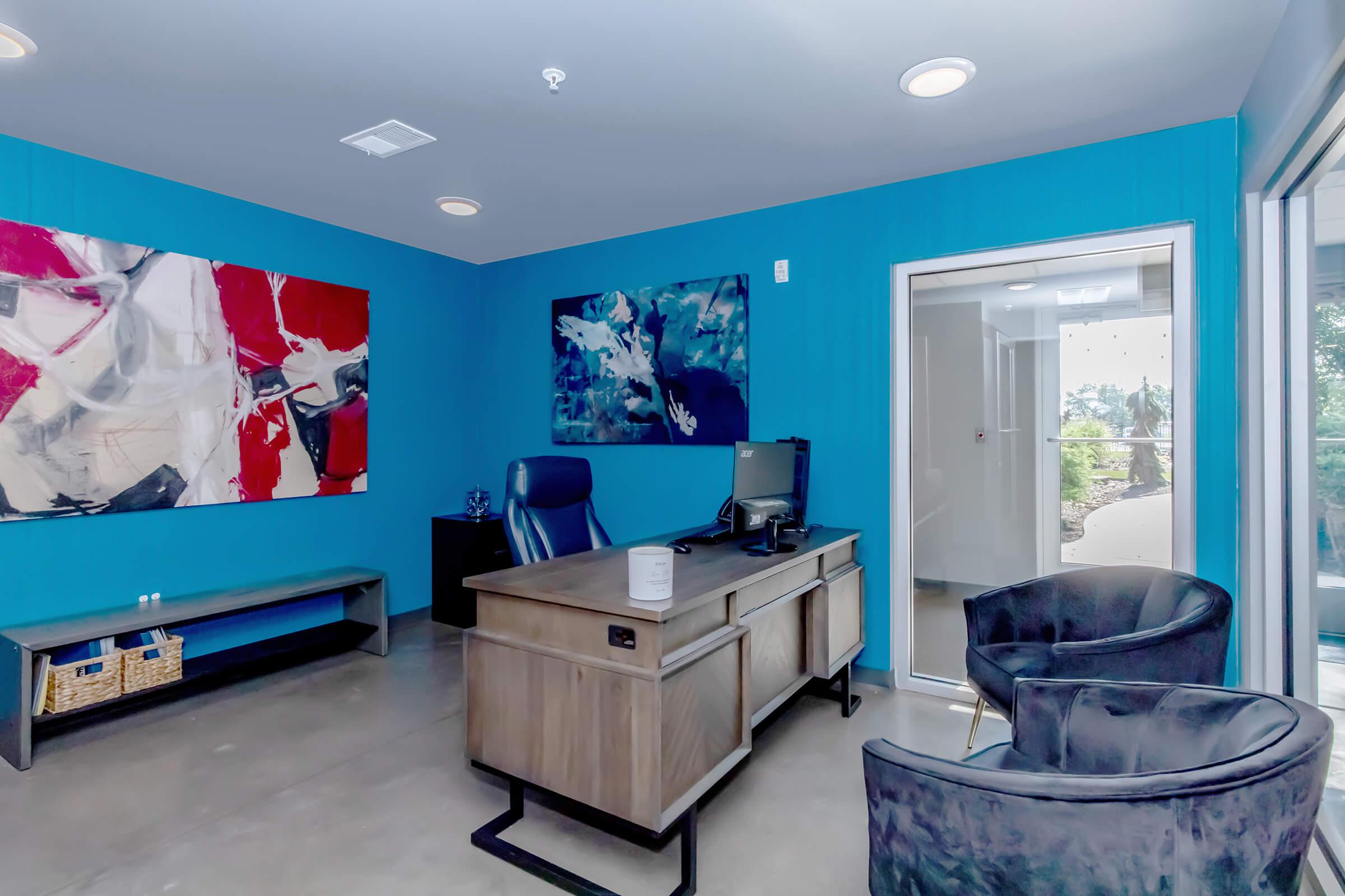 A modern office space featuring a blue accent wall, a wooden desk with a computer, and two stylish chairs. Abstract artwork adorns the walls, adding a pop of color. Large windows allow natural light to illuminate the room, creating a bright and inviting atmosphere. A sleek storage basket is visible beside the desk.