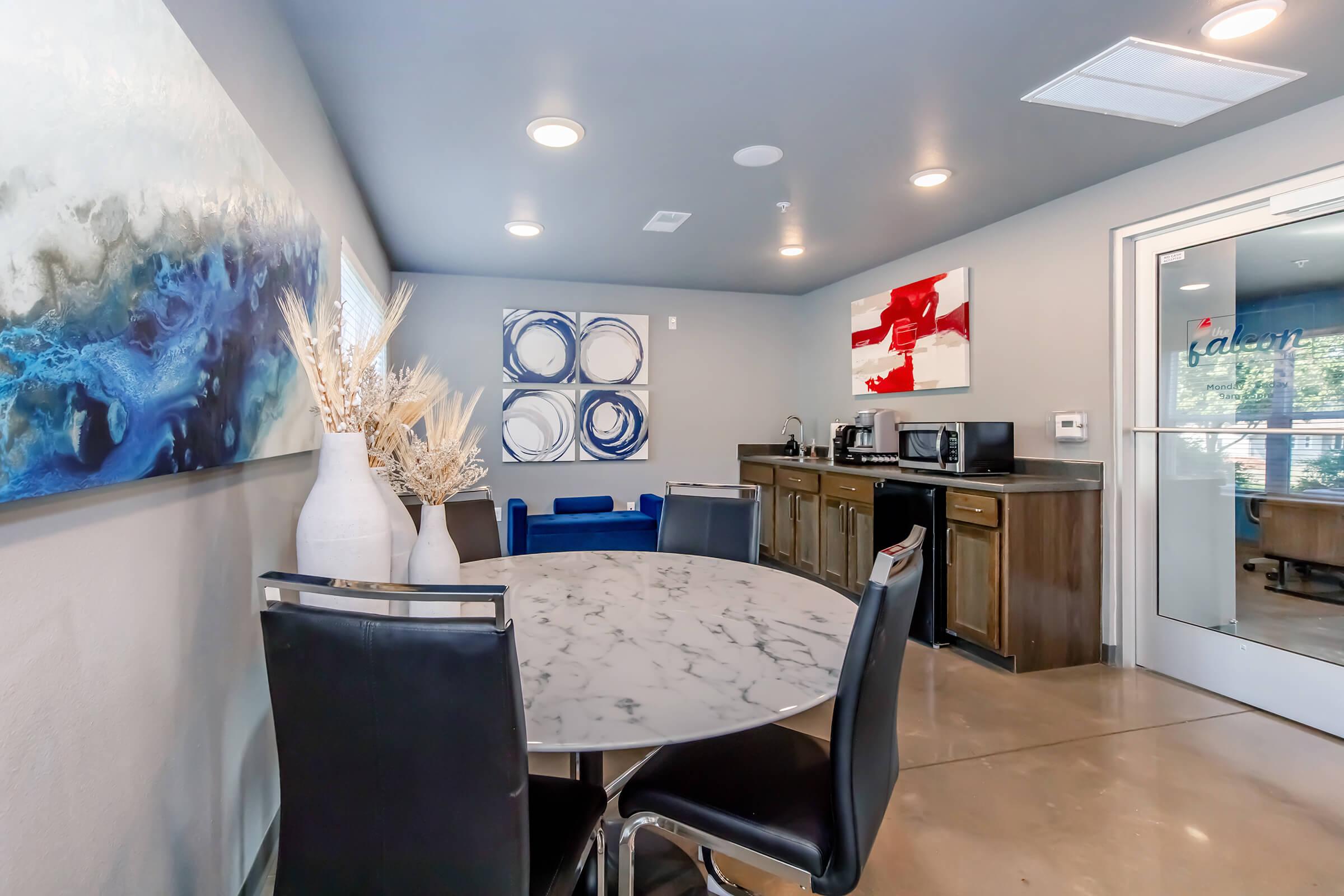 A modern communal space featuring a round marble table with black chairs. The room is decorated with abstract wall art, including blue and red pieces. There is a kitchenette with a microwave and coffee maker in the background, and a blue couch is visible to the side. The space is well-lit and contemporary.