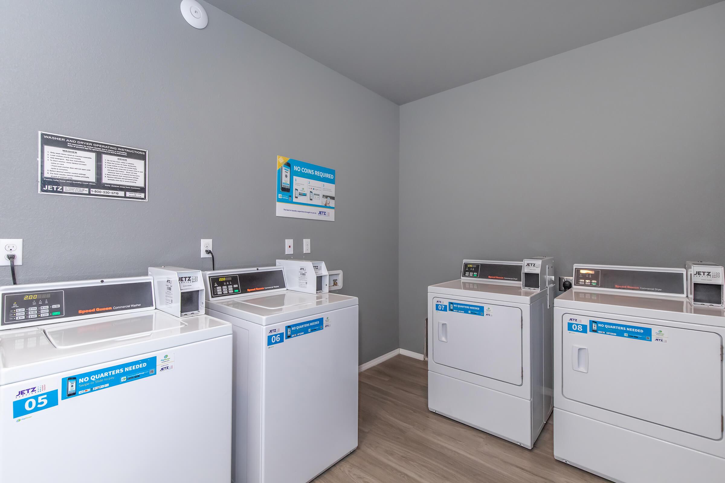 Laundry room featuring multiple coin-operated washer and dryer machines, all labeled with numbers 05 and 08. The machines are situated against a grey wall, with informational signs and operating instructions displayed. The floor is light-colored and the room is well-lit.