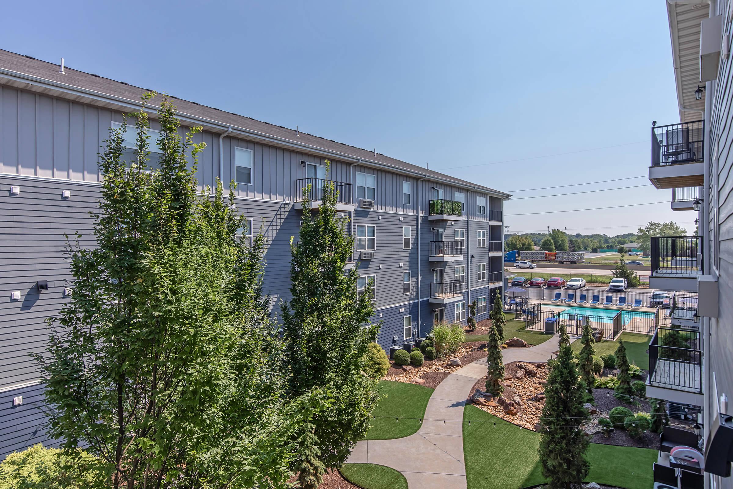 View of a modern apartment complex featuring a well-maintained courtyard with landscaping and swimming pool. The buildings are multi-story with gray siding, and there are cars parked nearby. Clear blue sky overhead enhances the inviting atmosphere of the residential area.