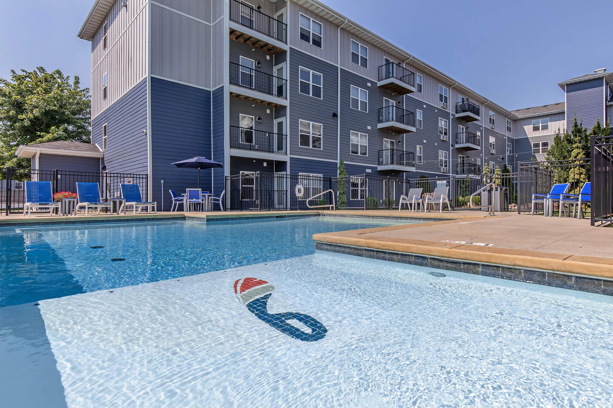 A clear swimming pool in the foreground with a colorful design on the bottom, surrounded by lounge chairs and umbrellas. In the background, a modern apartment building features balconies and multiple stories. The sky is bright blue, indicating sunny weather.