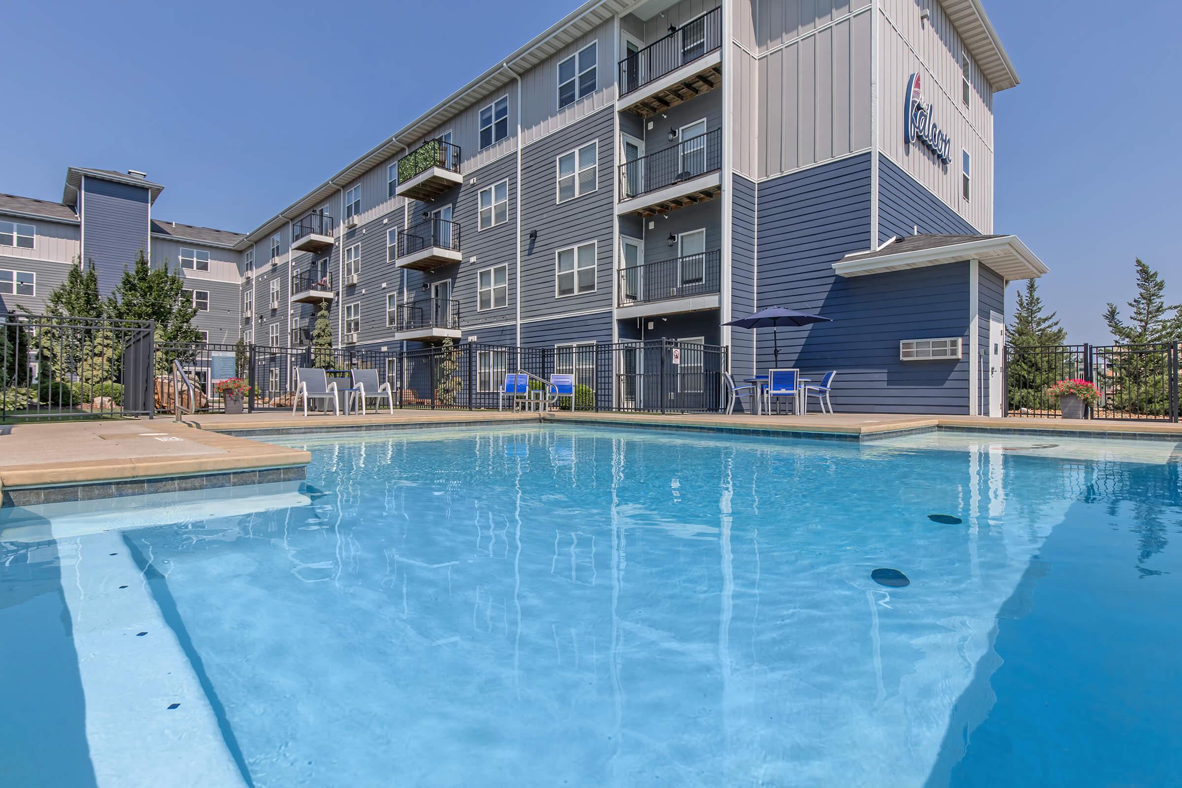 View of a swimming pool in front of a modern multi-story apartment building. The pool features clear blue water, with lounge chairs and umbrellas nearby. The building has a blue exterior with balconies, and plants enhance the outdoor area. Clear skies are visible above, indicating a sunny day.