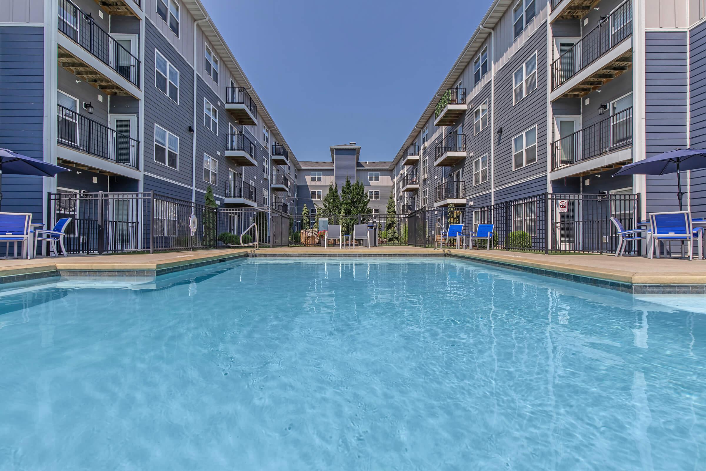 A clear swimming pool reflecting blue skies, surrounded by modern apartment buildings with balconies. Lounge chairs are set up around the pool area, creating a relaxing atmosphere. Lush greenery is visible in the background, enhancing the serene space.