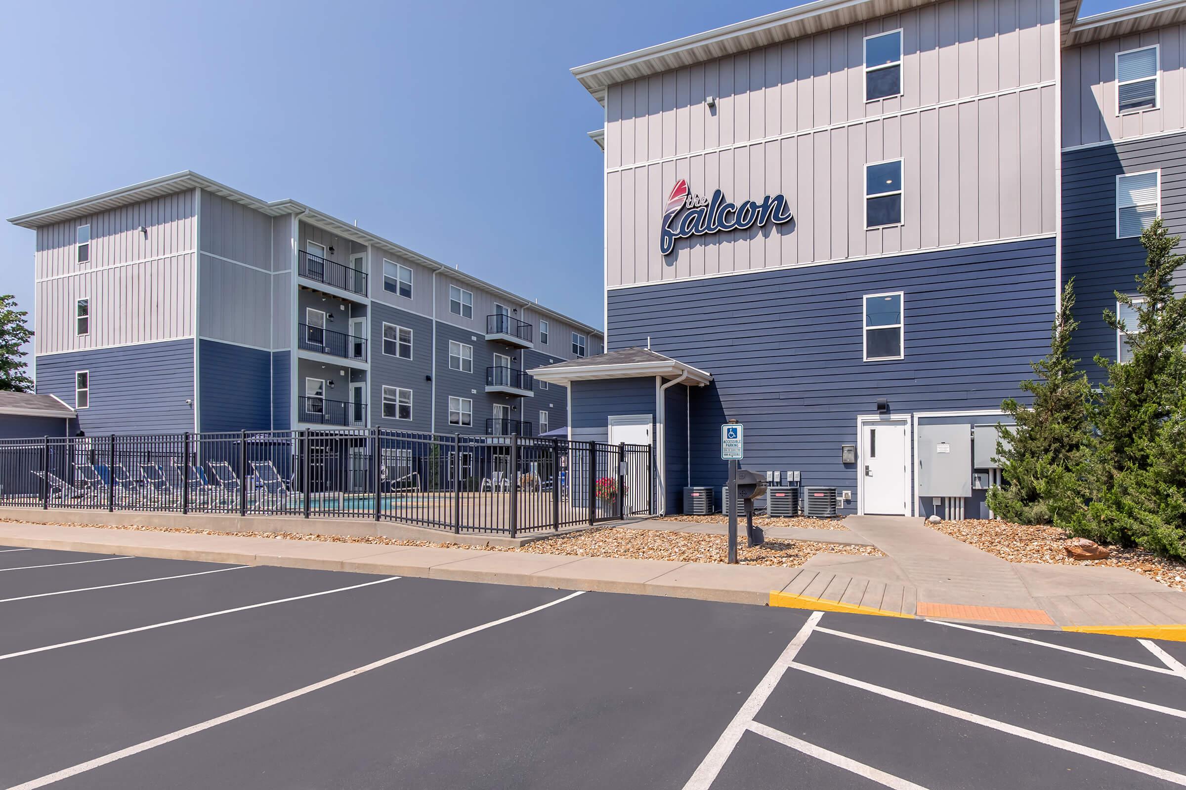 Exterior view of a modern apartment complex featuring two buildings with blue and gray siding. In the foreground, a paved parking area is visible. The complex includes a fenced pool area with lounge chairs, and the name “Falcon” is prominently displayed on one of the buildings. Clear blue sky above.