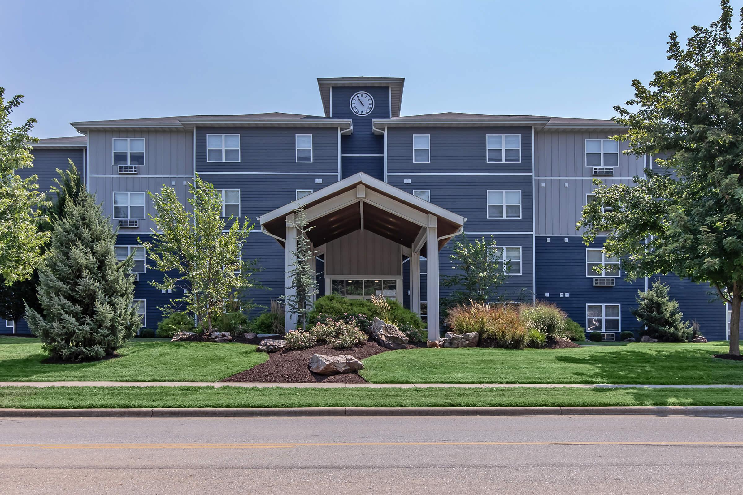 A modern multi-story apartment building with a clock tower feature. The entrance is covered by a sheltered portico, surrounded by landscaped greenery, including trees and shrubs, set on a clear blue sky day. The scene includes a well-maintained lawn and decorative rocks, providing a welcoming appearance.