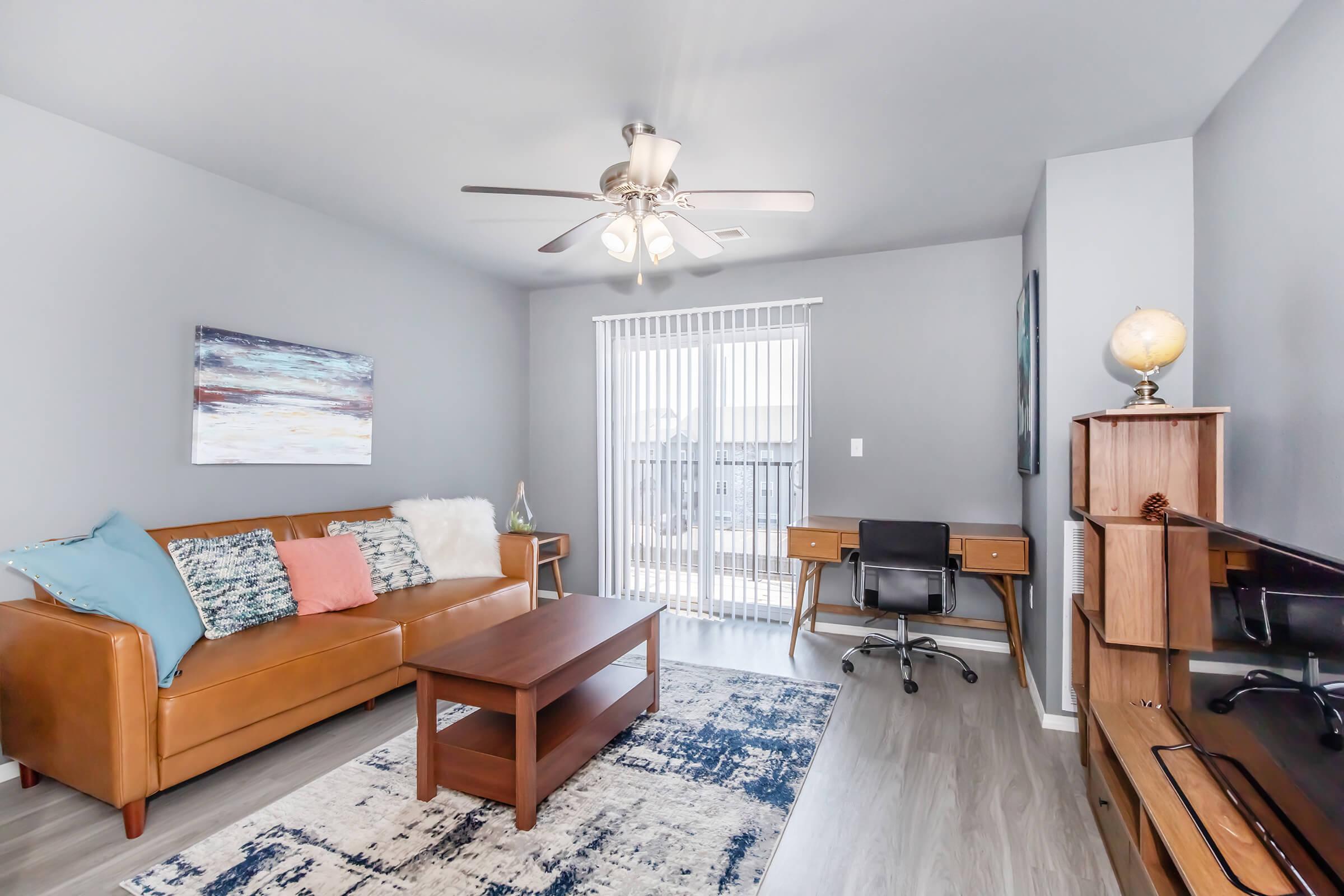 A cozy living room featuring a brown leather sofa with colorful cushions, a wooden coffee table, a modern desk with a chair, and a TV. The walls are painted grey, and large windows with vertical blinds allow natural light. Decorative items include a globe and wall art. The flooring is light wood.