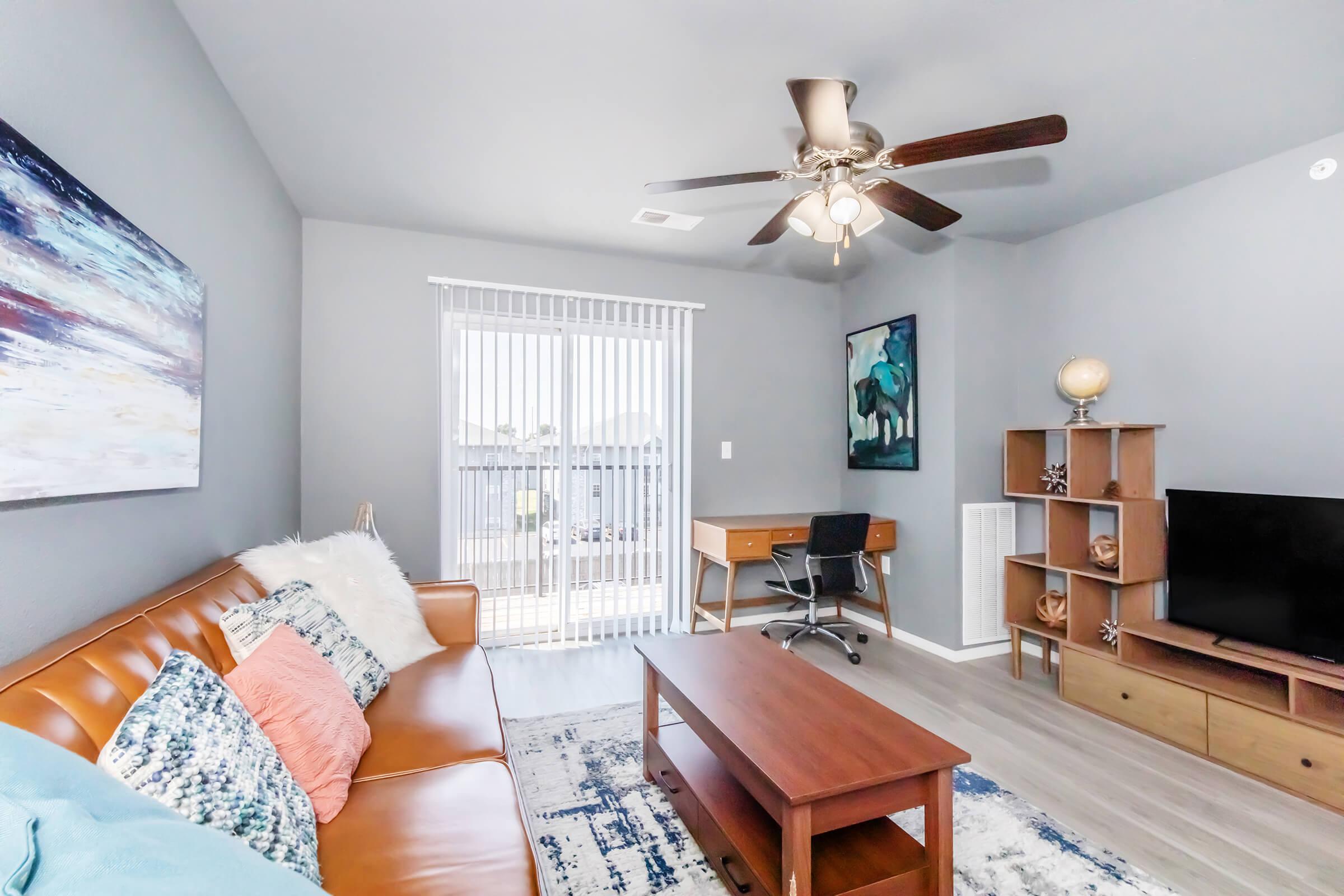 Modern living room featuring a brown leather sofa with decorative pillows, a wooden coffee table, and a desk with an office chair. A large window with vertical blinds allows natural light, and a TV unit is displayed against the wall. Art and decor items add a personal touch to the space.