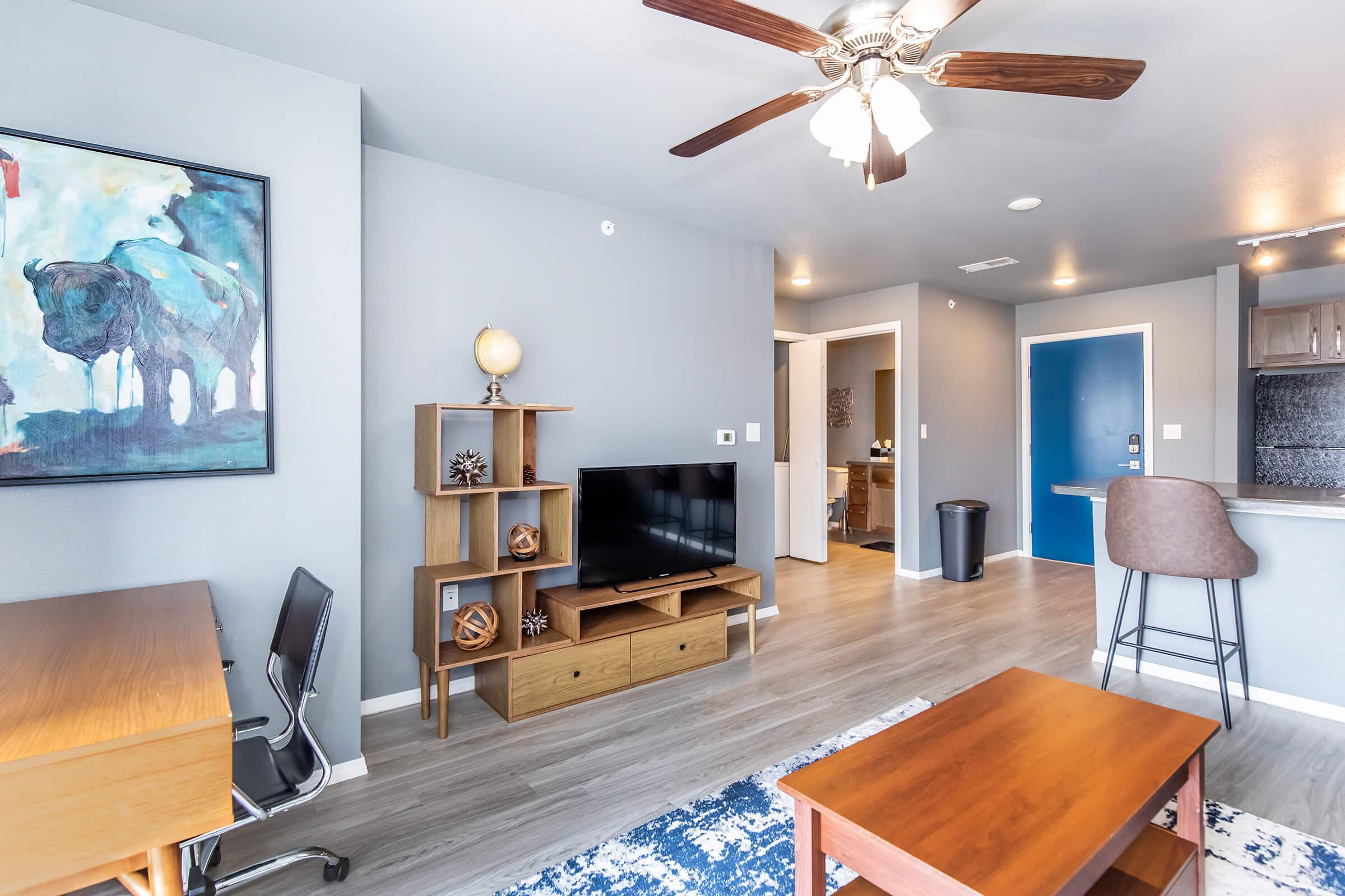 A modern living room with light gray walls featuring a wooden coffee table, a TV stand with a television, and a desk with a chair. A ceiling fan is overhead, and there’s a blue front door visible. The room has an artistic painting of a buffalo on the wall and a small decorative globe on the shelf.