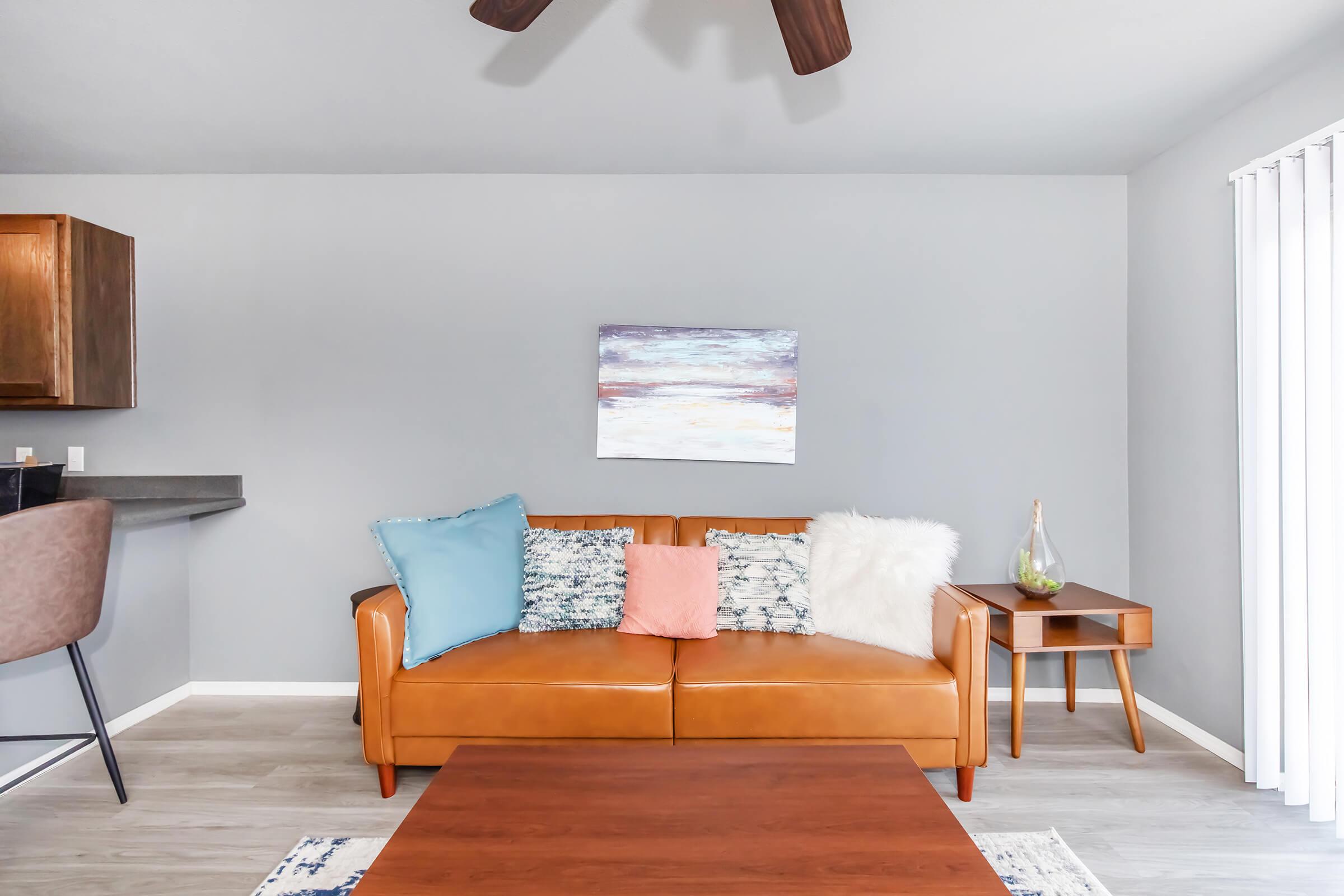 A modern living room featuring a brown leather sofa adorned with colorful pillows, a wooden coffee table, and a small side table with a decorative vase. The walls are painted light grey, and there is a piece of abstract artwork above the sofa. Natural light streams in through vertical blinds.