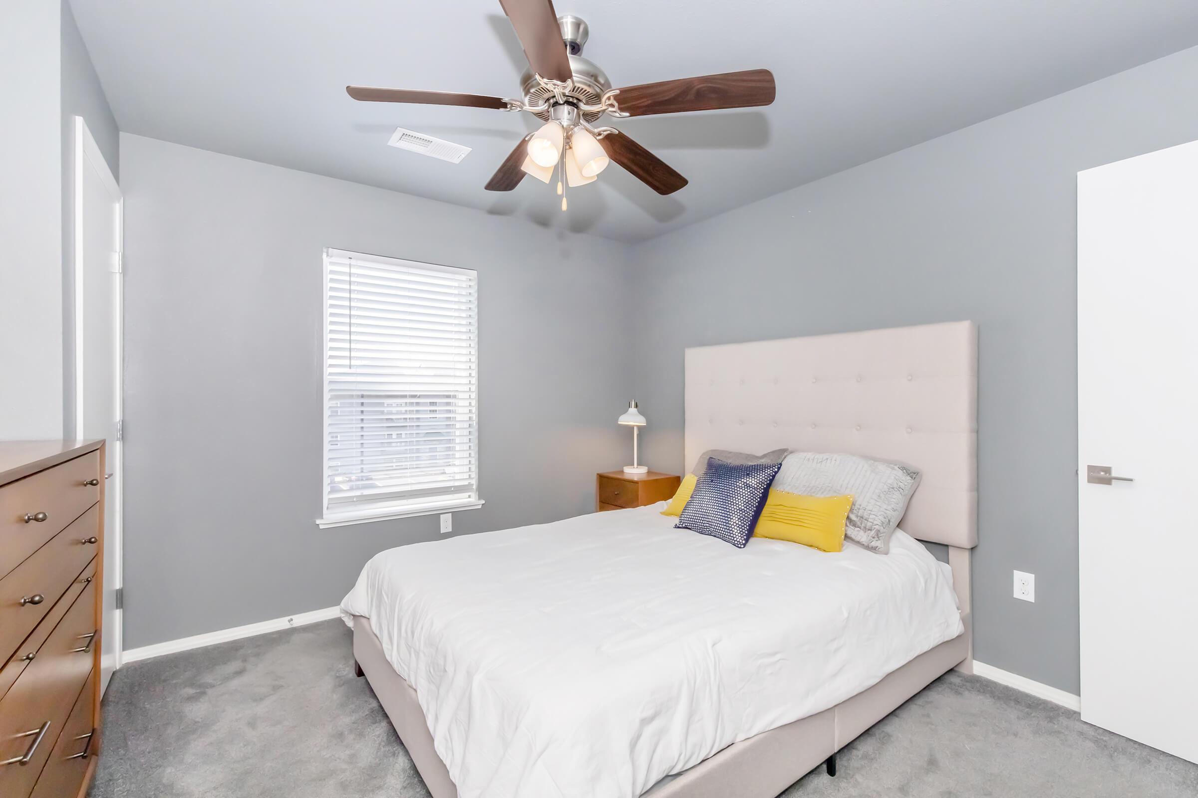 A cozy bedroom featuring a light gray wall, a queen-sized bed with a soft pink headboard and white bedding, accented by yellow and blue decorative pillows. A wooden nightstand holds a lamp next to the bed, and a dresser is visible on the left. A ceiling fan with wooden blades adds a rustic touch. Natural light streams in through the window.