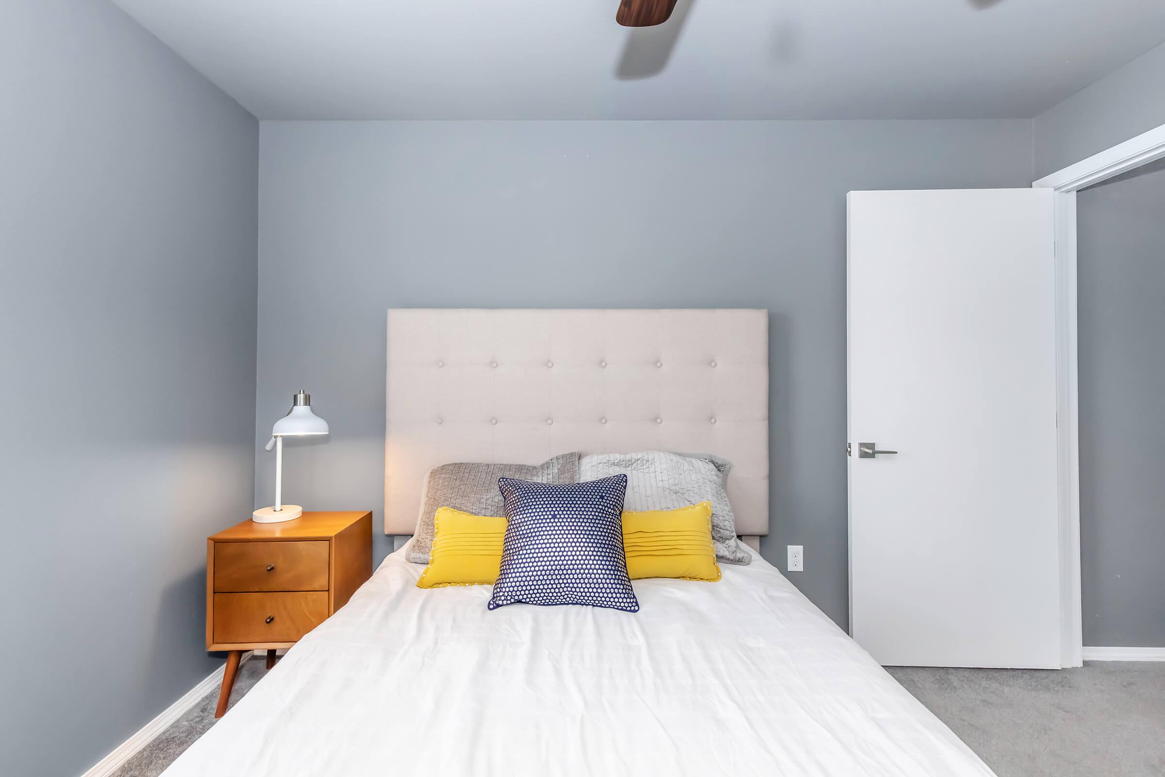 A modern bedroom featuring a light gray wall, a beige upholstered headboard, a white bed with a gray and blue patterned blanket, and two yellow throw pillows. A bedside table with a lamp is on one side, and a closed white door is visible in the background. The overall decor is simple and contemporary.