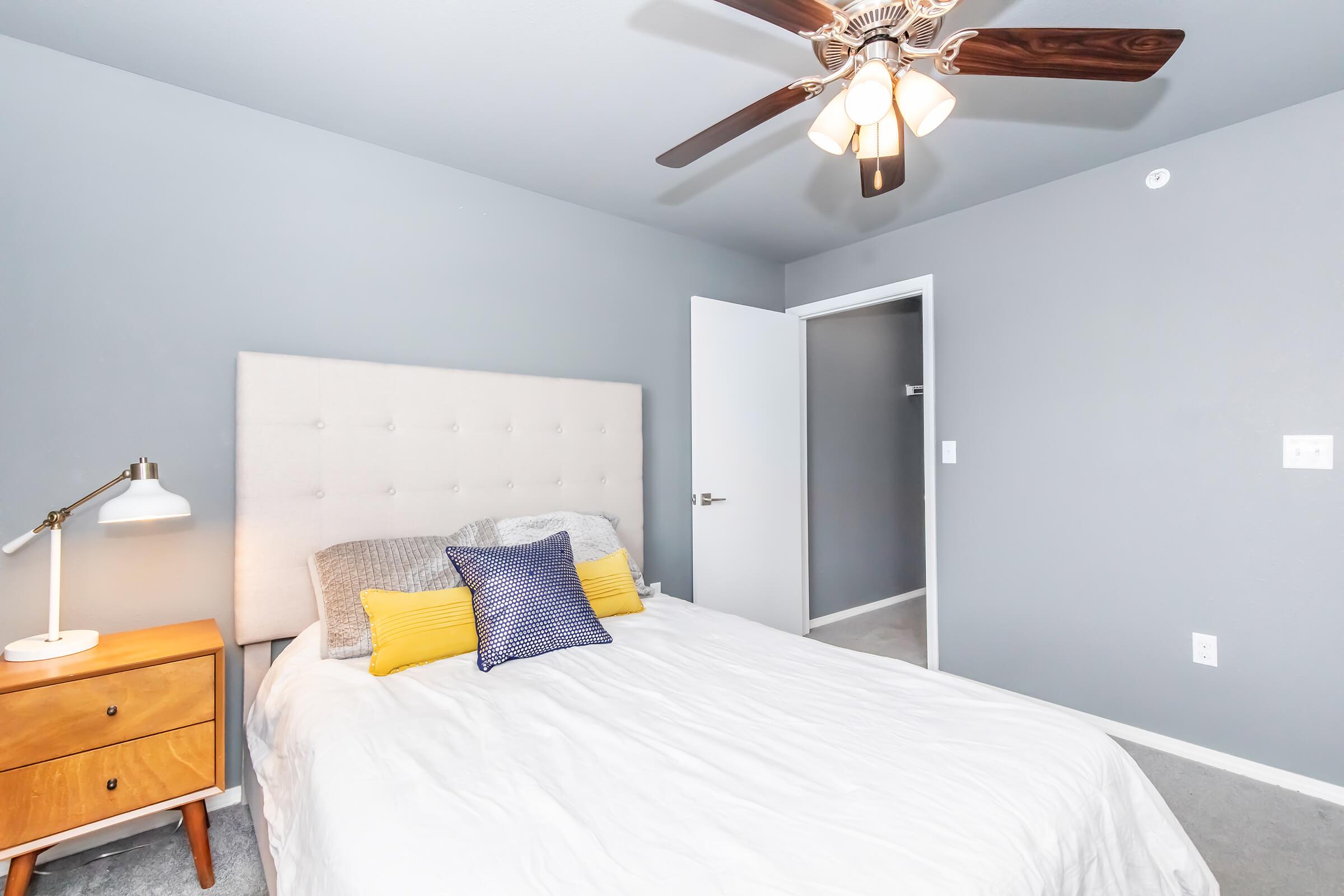 A modern bedroom featuring a queen-sized bed with light bedding and decorative pillows. There is a wooden nightstand with a lamp beside the bed. The walls are painted gray, and a ceiling fan with five blades hangs above. A closet door is visible in the background, and the flooring is carpeted.