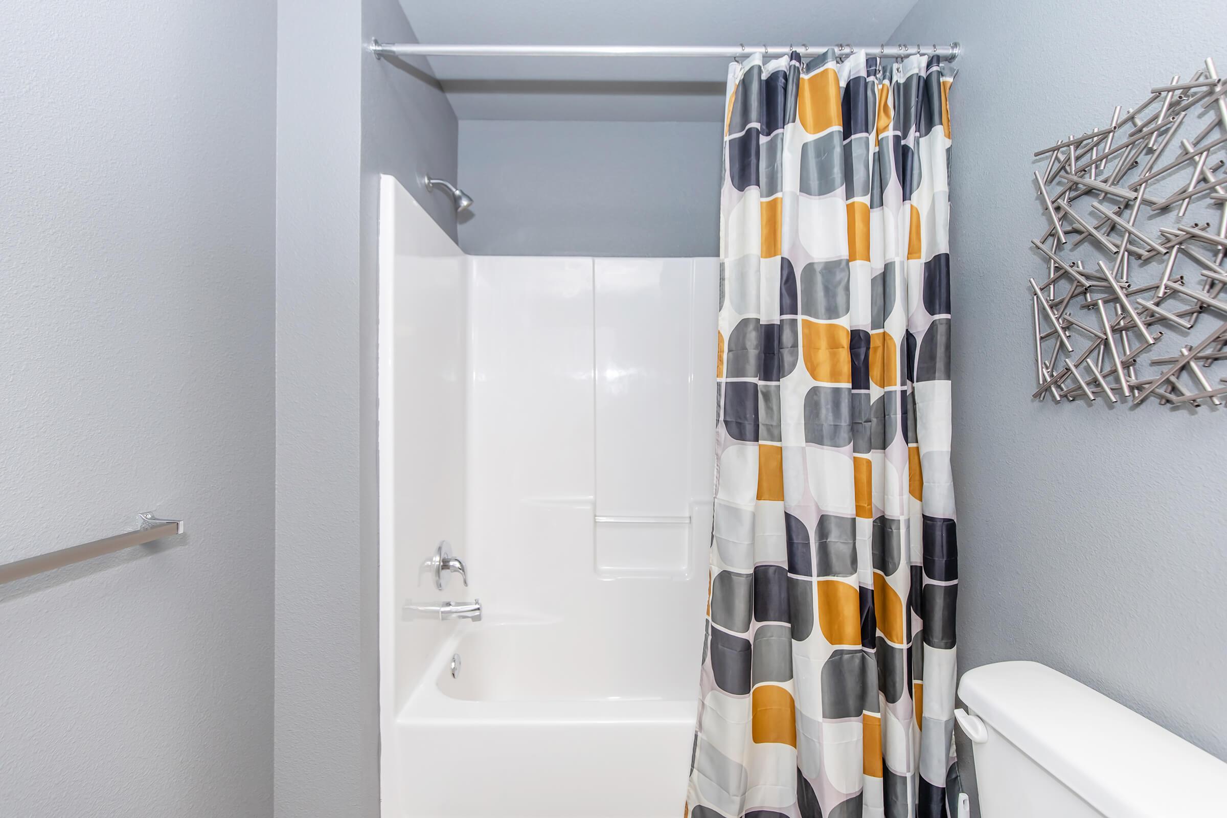 A modern bathroom featuring a white bathtub and shower, a colorful geometric-patterned shower curtain in shades of yellow, gray, and white, and a sleek toilet. The walls are a light gray, and there's a decorative wall piece made of metal rods.